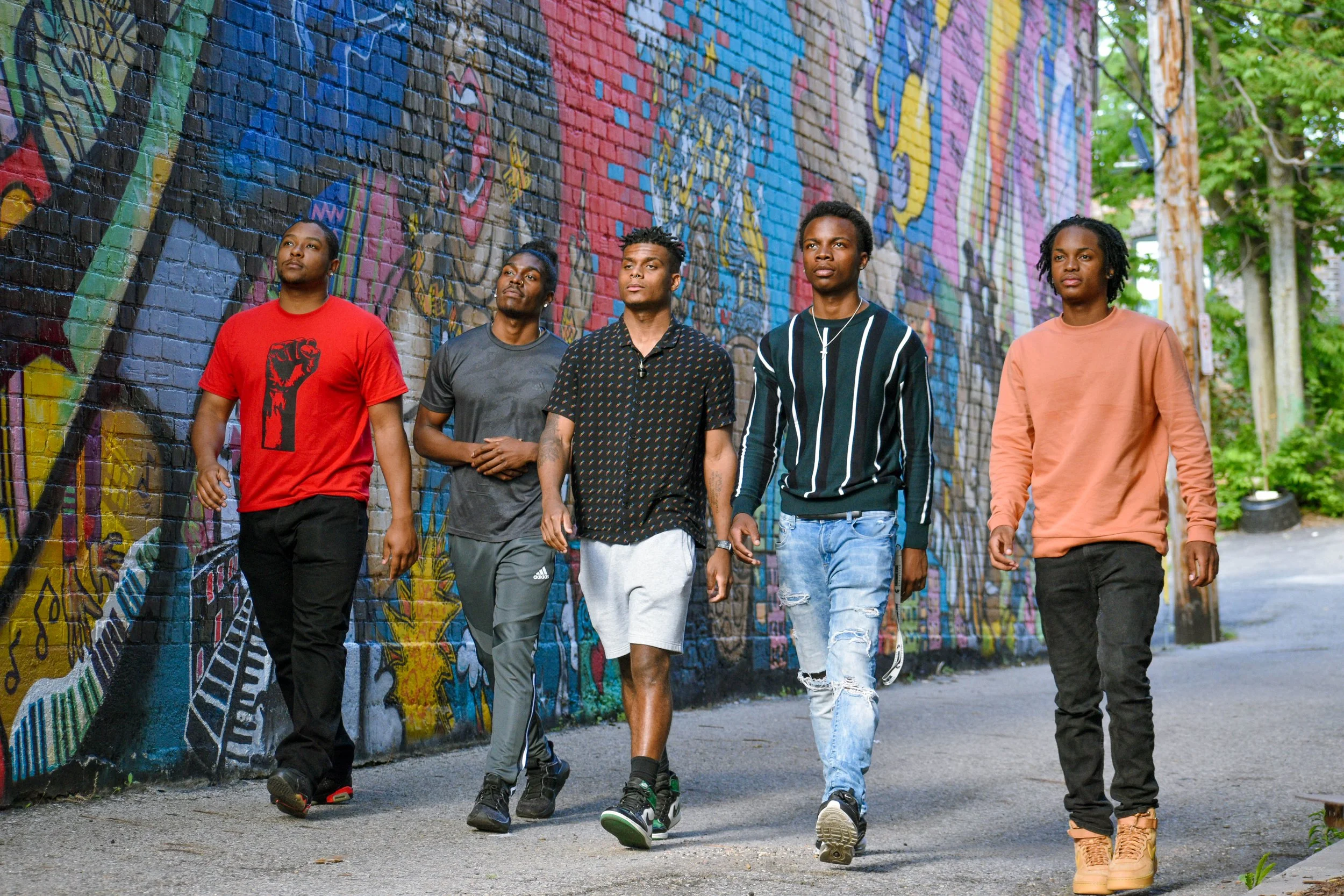 Five young men walking together along a city street with a colorful graffiti mural on a brick wall behind them, surrounded by green trees.