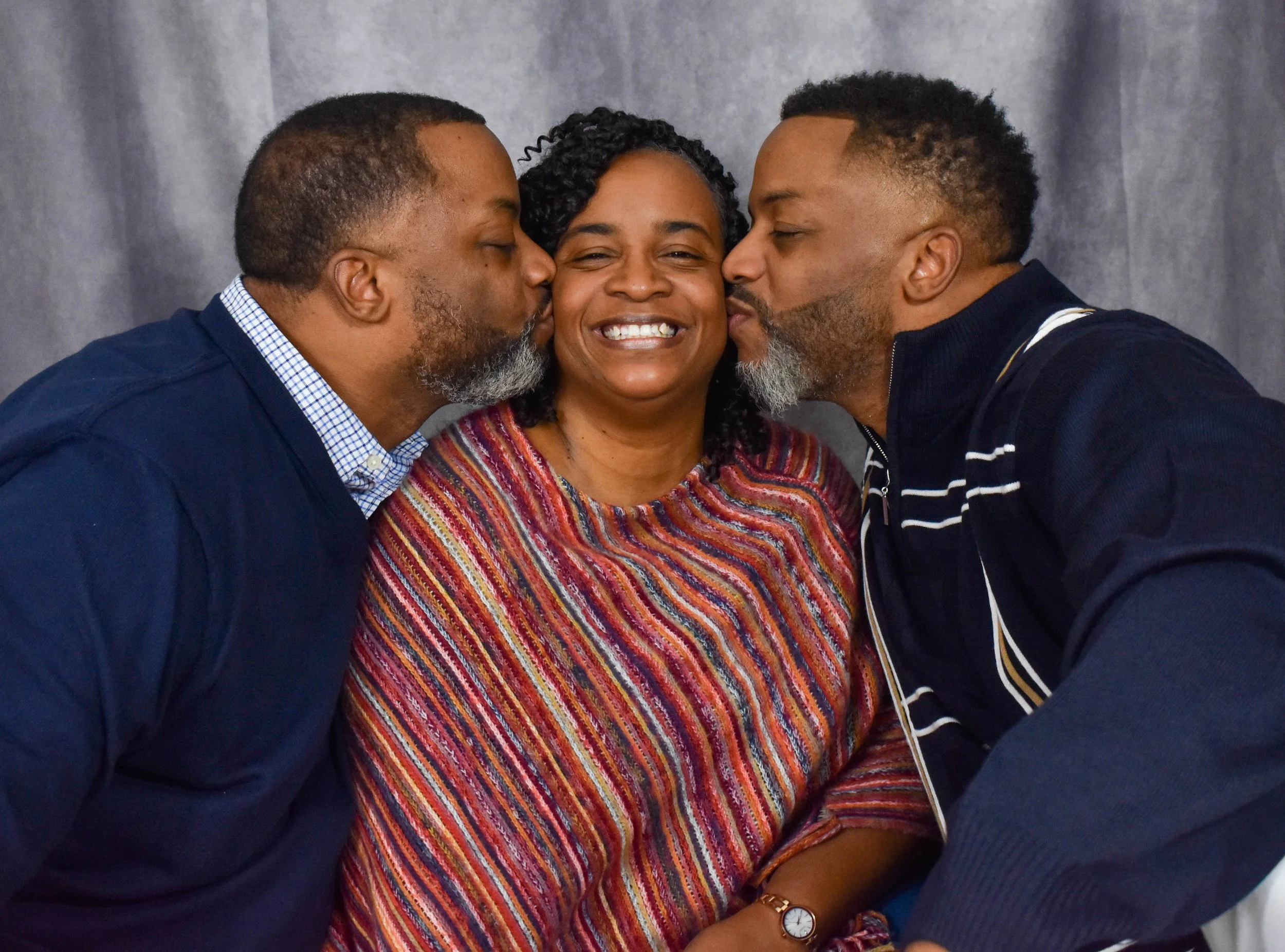 Two men kissing a woman on each cheek, with her smiling in the middle. They are all close together and have their eyes closed, against a gray background.