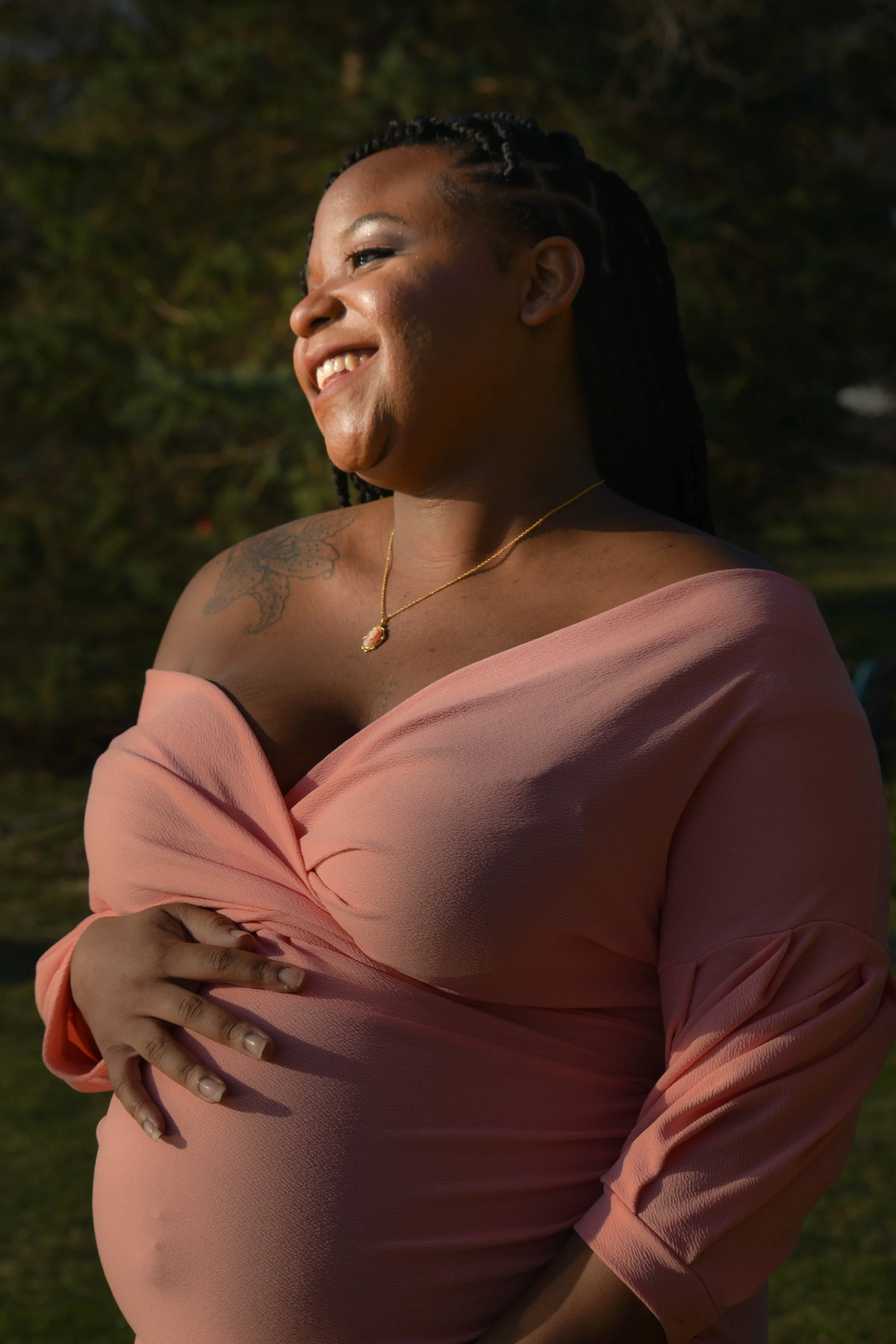 A smiling black woman with braided hair wearing a pink off-shoulder dress and a gold necklace, standing outdoors with greenery in the background.