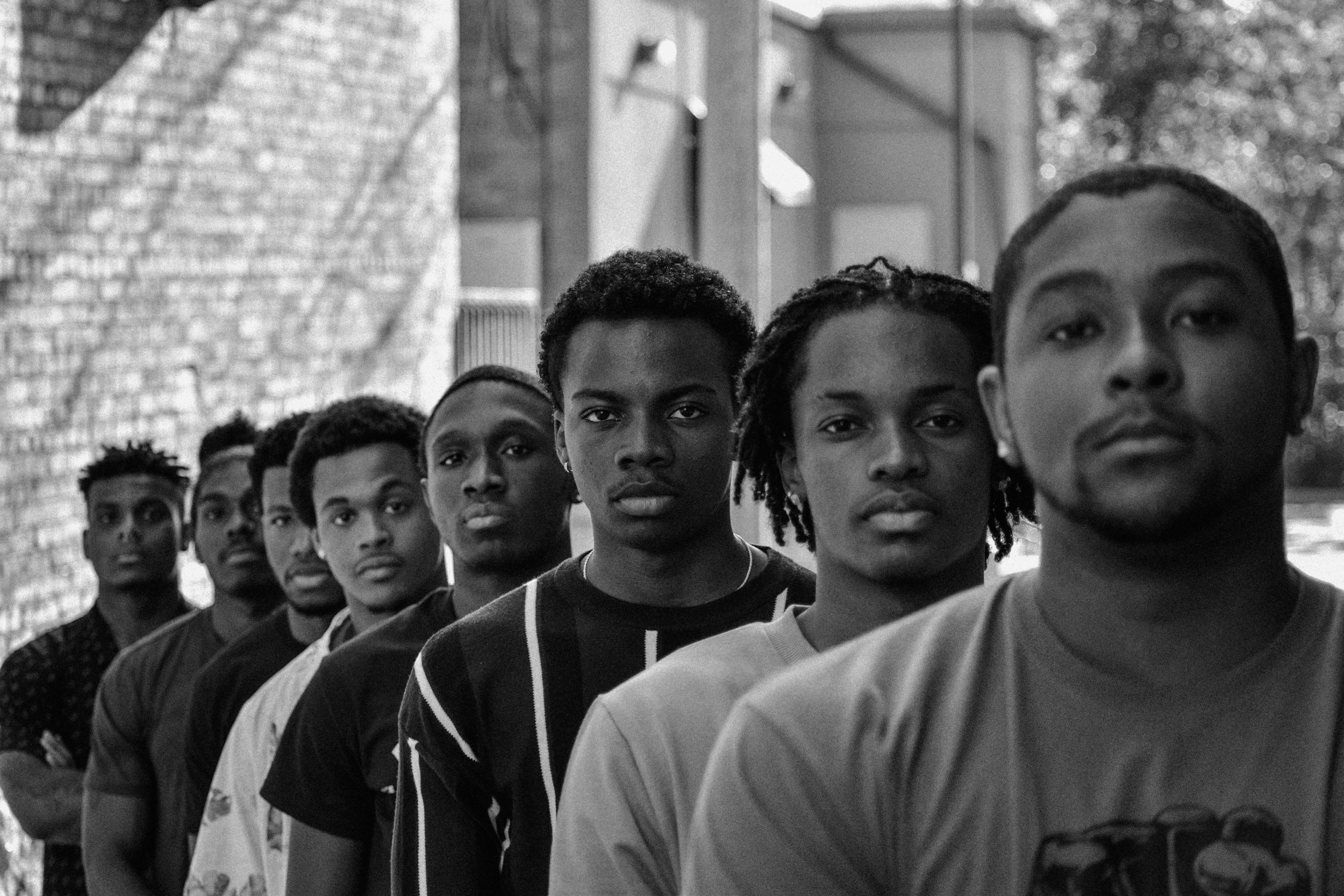 A line of young people standing outdoors in an urban setting, all looking directly at the camera. The image is in black and white, with a brick building in the background.