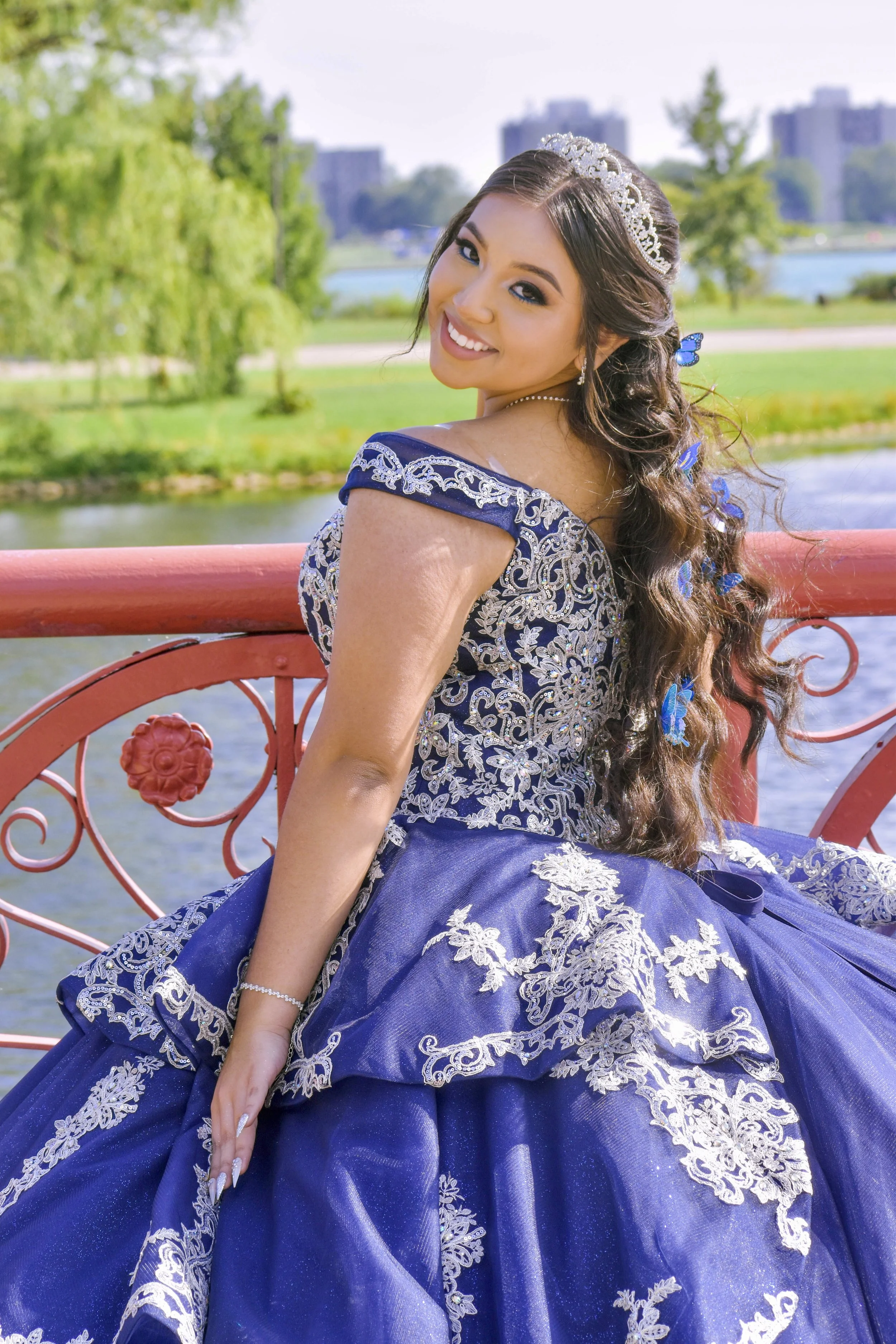 A young woman in a blue and silver gown with intricate embroidery, wearing a tiara and jewelry, smiling and sitting on a red decorative bench outdoors near a river with greenery and city buildings in the background.