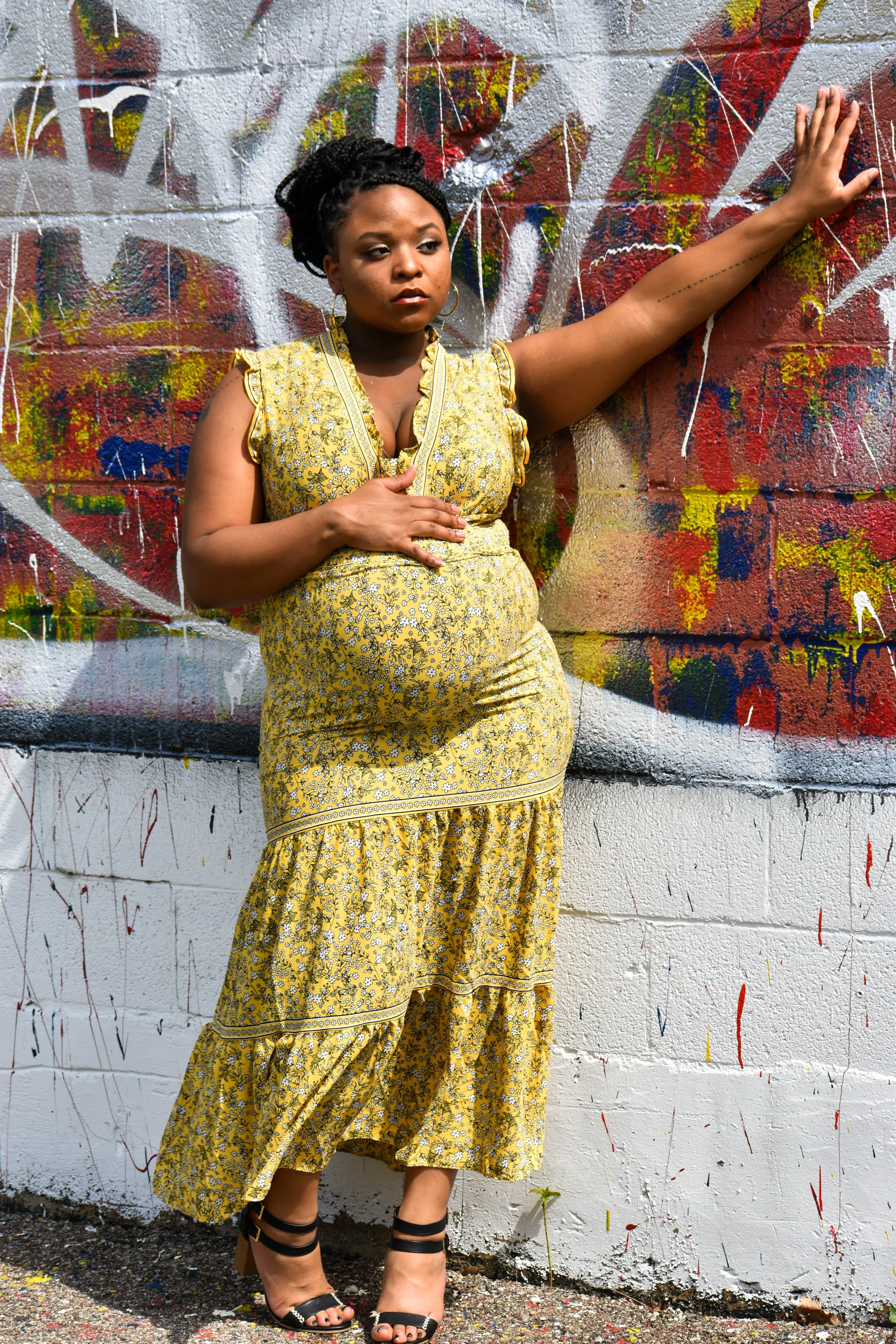 A woman wearing a yellow floral dress and black strappy heels standing against a colorful graffiti wall with her left arm extended and resting on the wall.