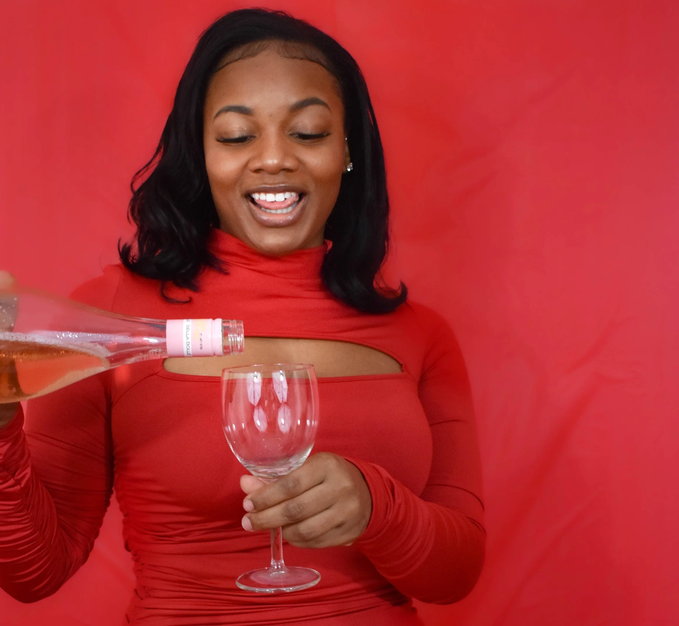 A woman with dark hair and jewelry, wearing a red top, is pouring a bottle of rosé wine into a glass against a red background.