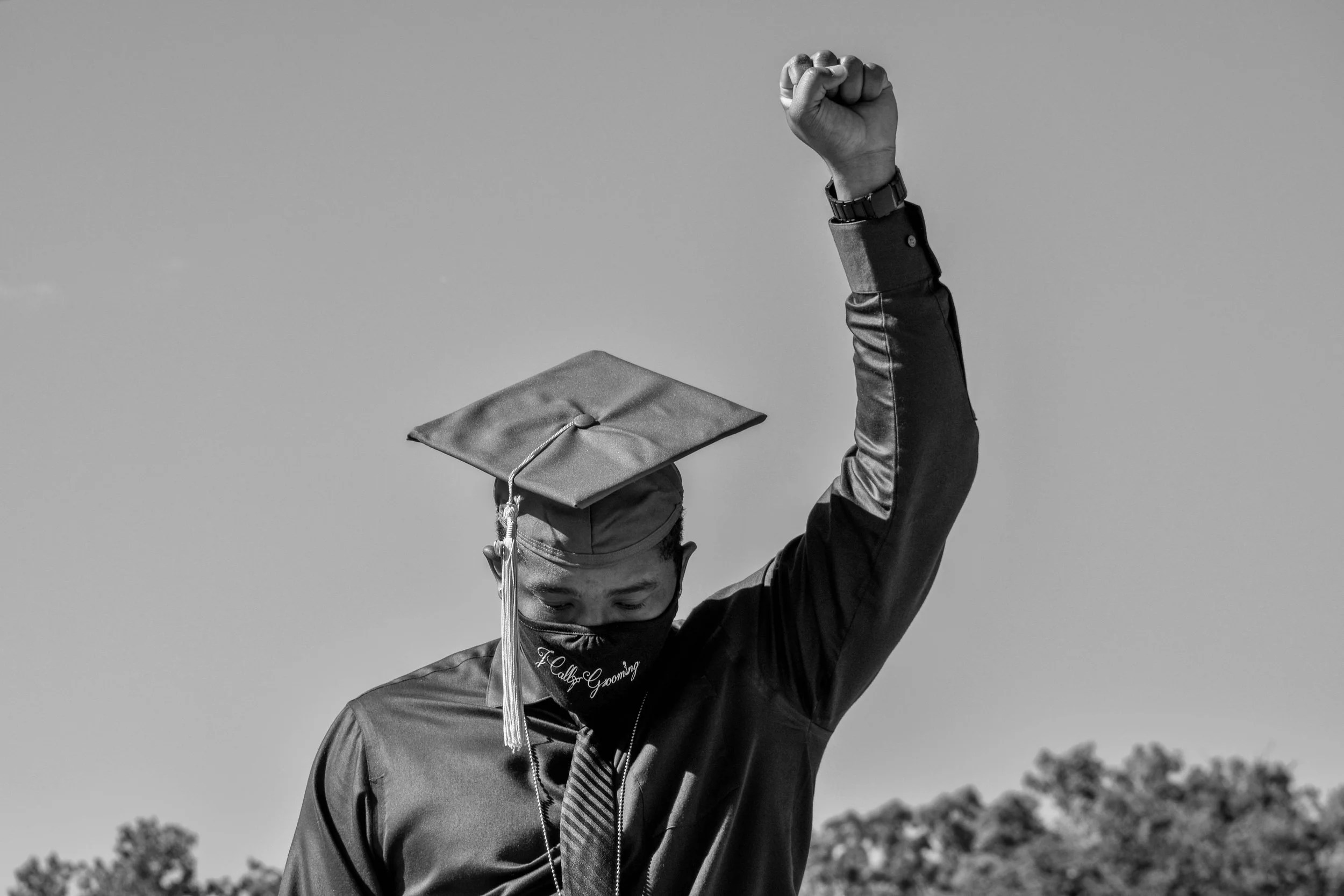 A graduate in a black gown and cap, with a tassel, raises one arm in celebration, holding the cap in the air during a graduation ceremony outdoors.