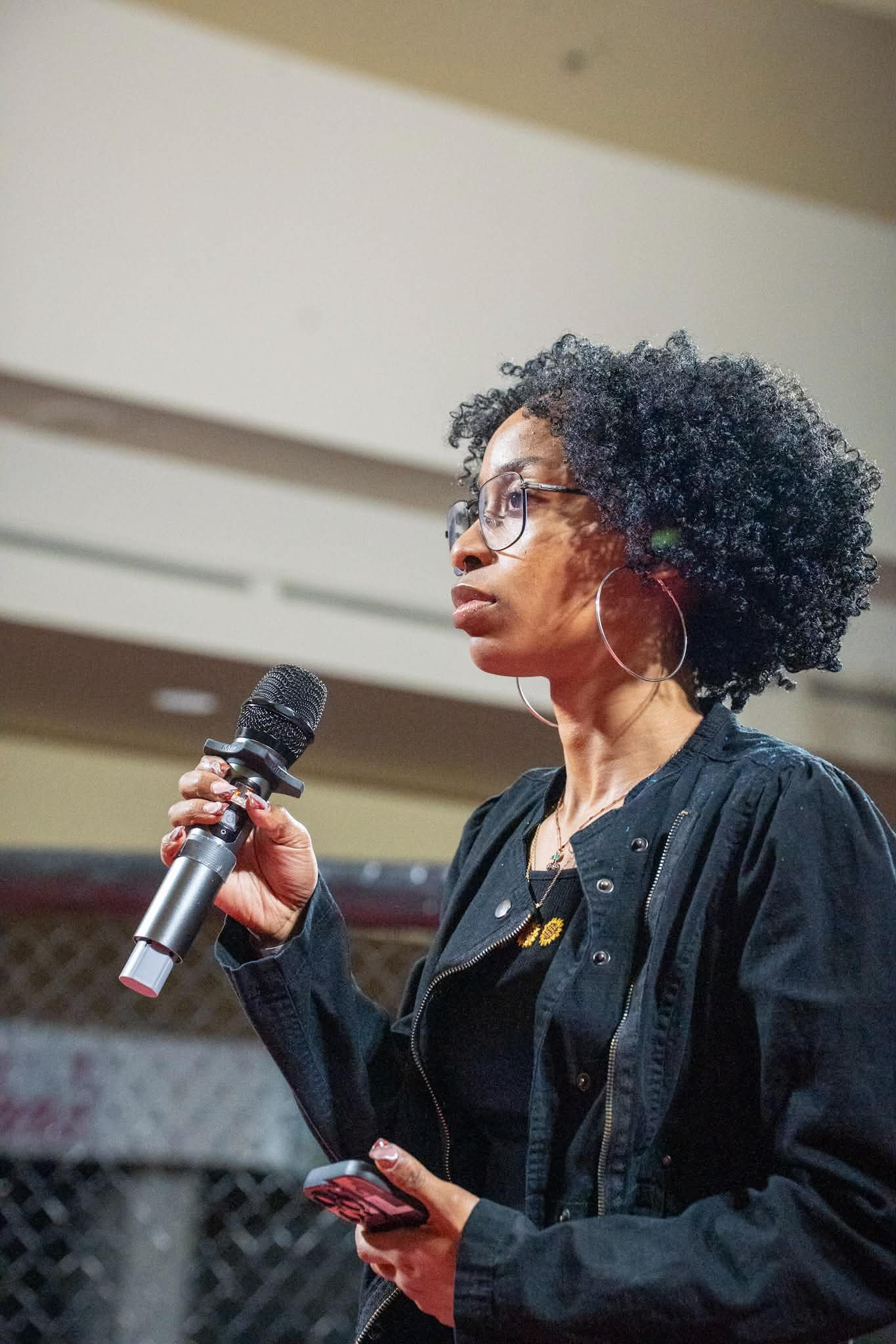A woman with curly black hair, glasses, and large hoop earrings holding a microphone and a smartphone, standing in an indoor setting.