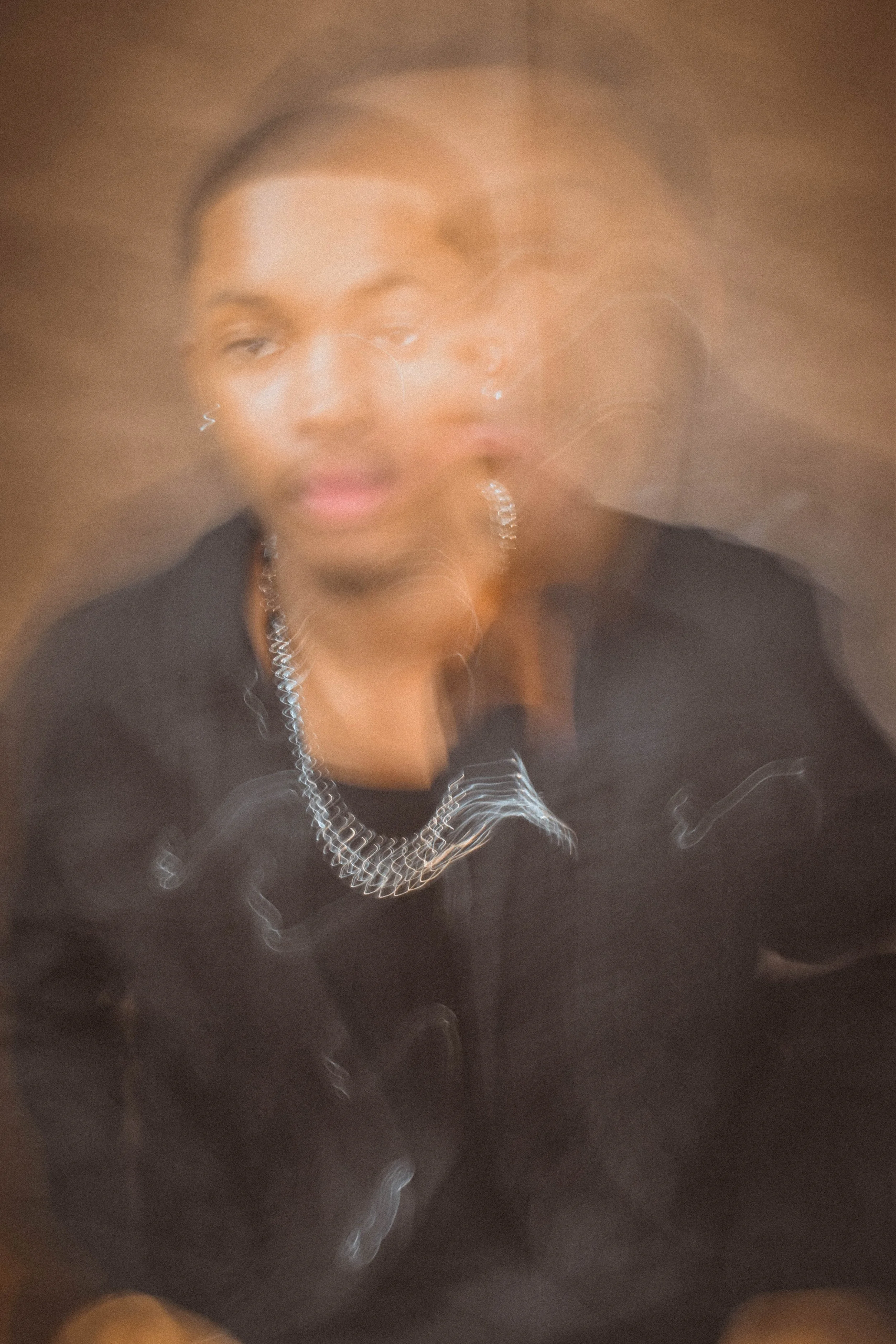 A person with dark hair and earrings, wearing a black top and a chain necklace, is in a space with blurred, swirling lights of various shapes and colors.
