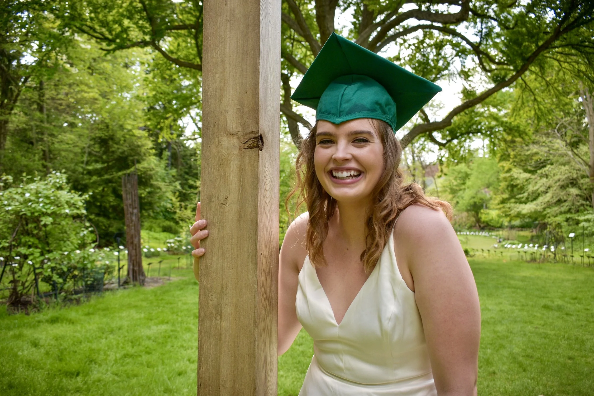 A young woman in a white dress and a green graduation cap holding a wooden post in a park with lush trees and grass.