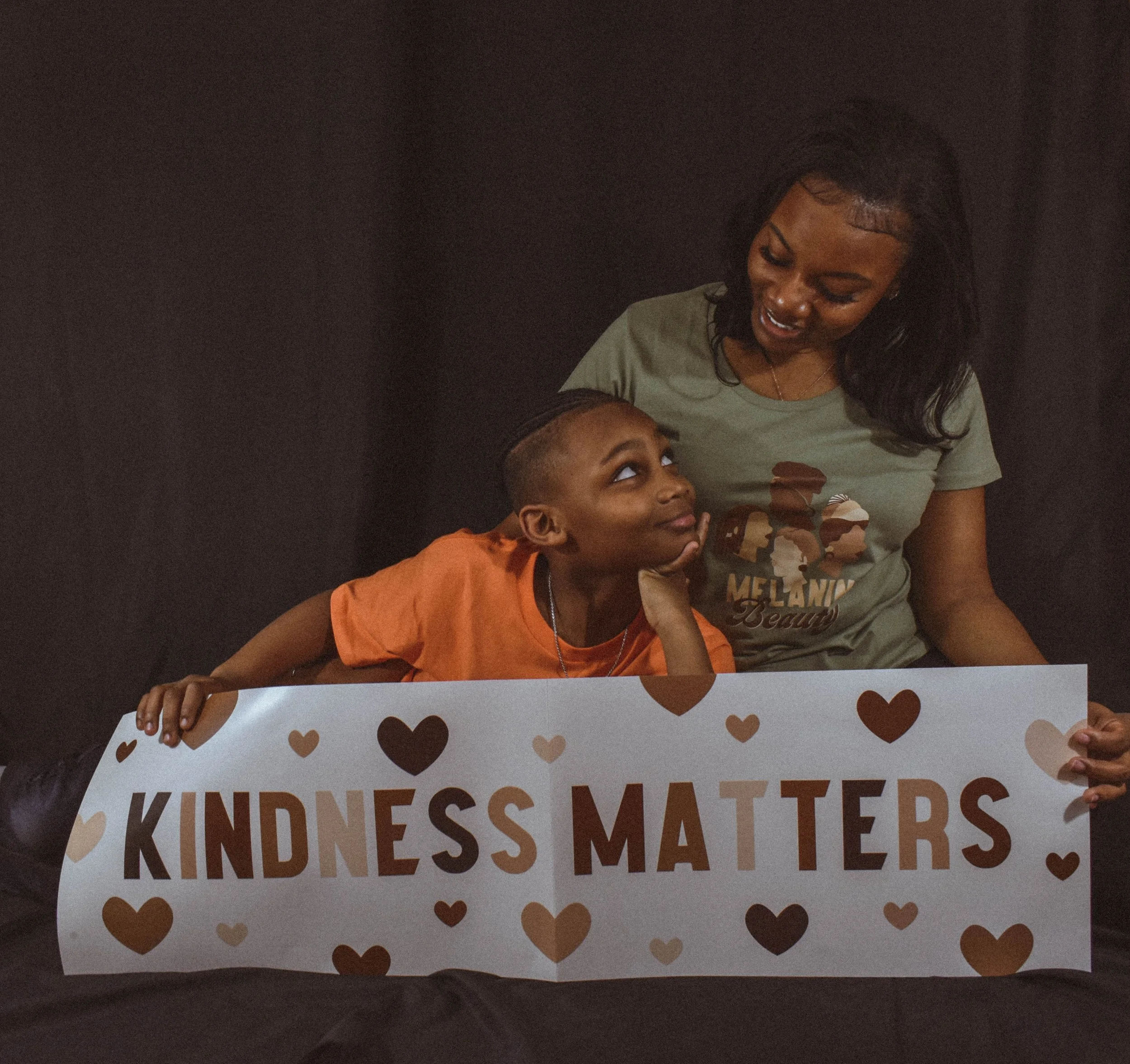 A woman and a young boy holding a sign that reads 'KINDNESS MATTERS' decorated with heart shapes, against a dark background.