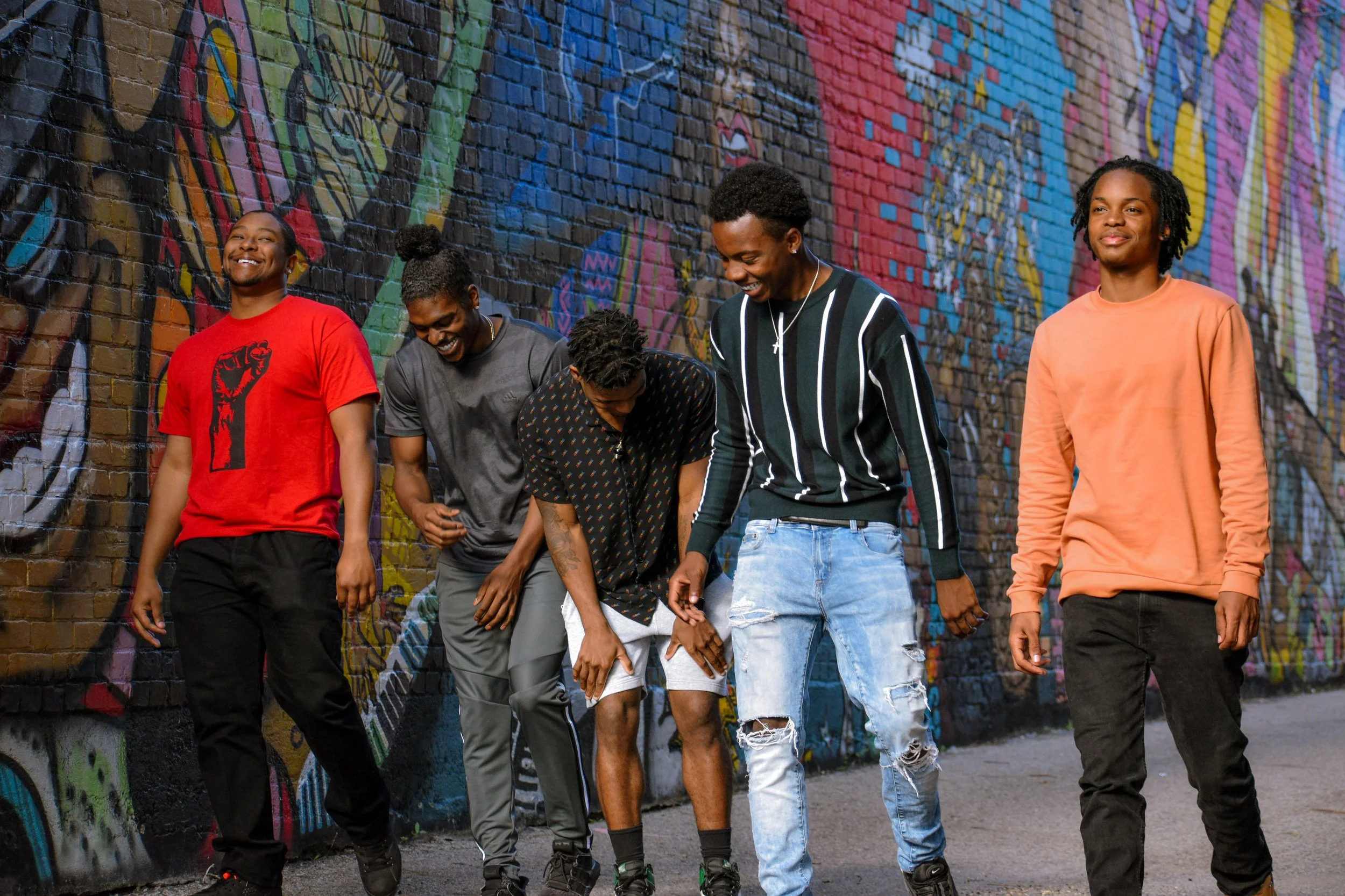 A group of five young men walking near a colorful graffiti wall in an urban setting. They are smiling and appear to be enjoying themselves.