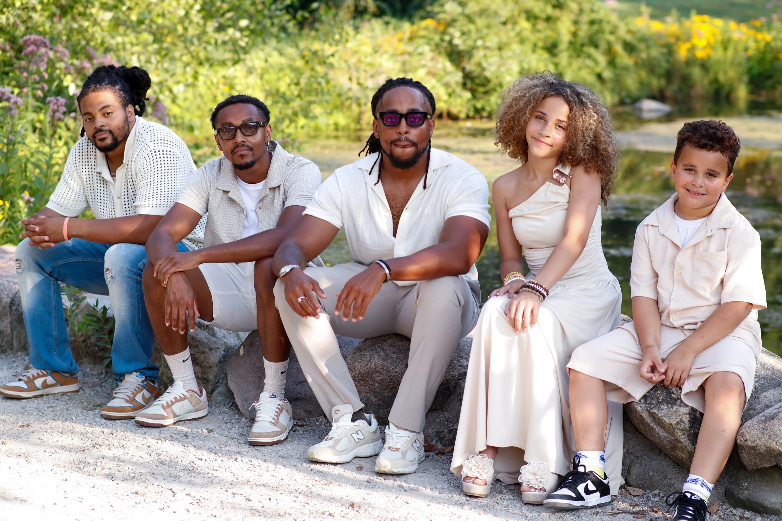 A diverse group of six people sitting on a rock by a river in a park, smiling and relaxing on a sunny day with greenery in the background.