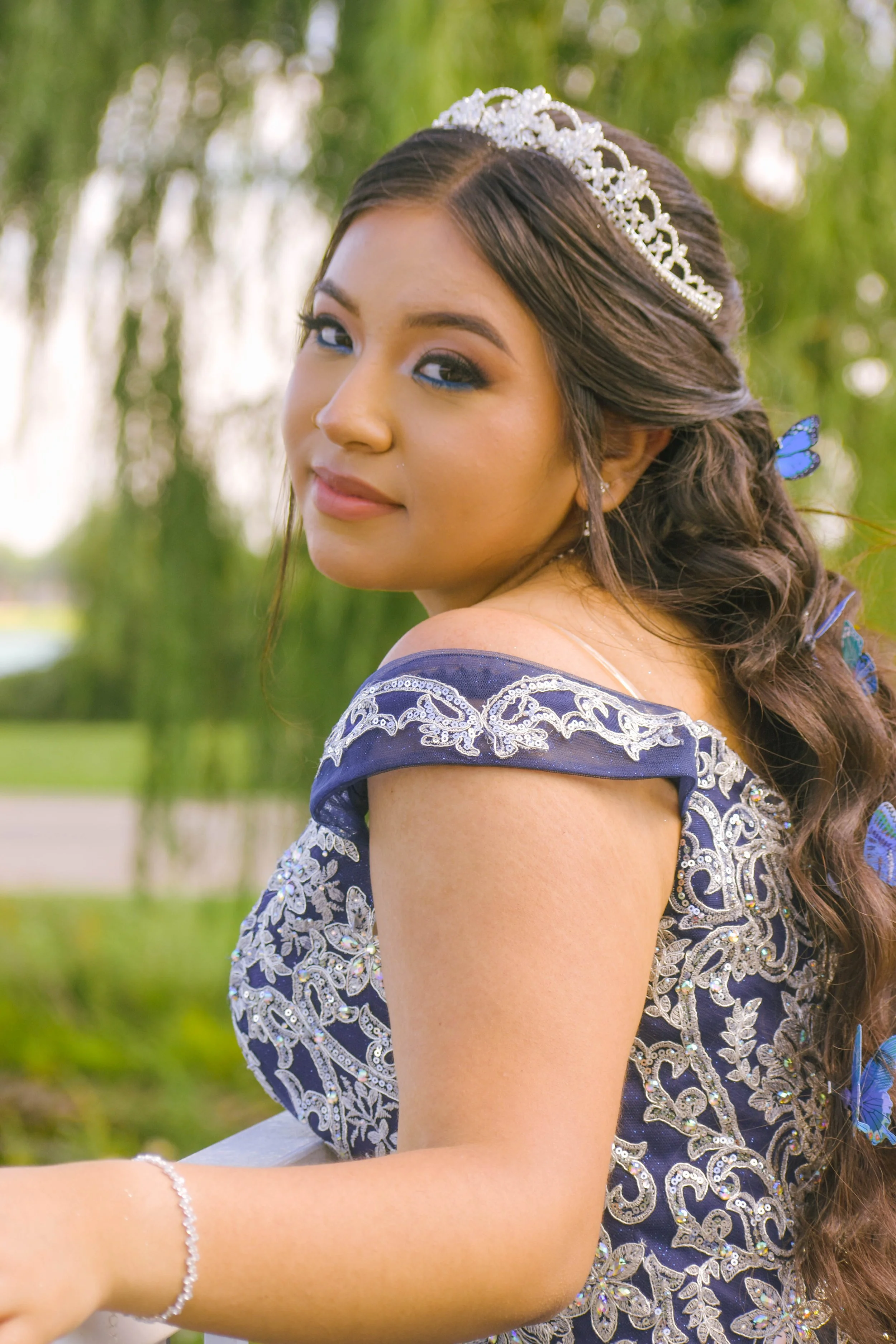 A young woman with brown hair styled in loose curls, wearing a silver tiara and a dark blue dress with silver embroidery, stands outdoors with a blurred green background, looking over her shoulder with a slight smile.