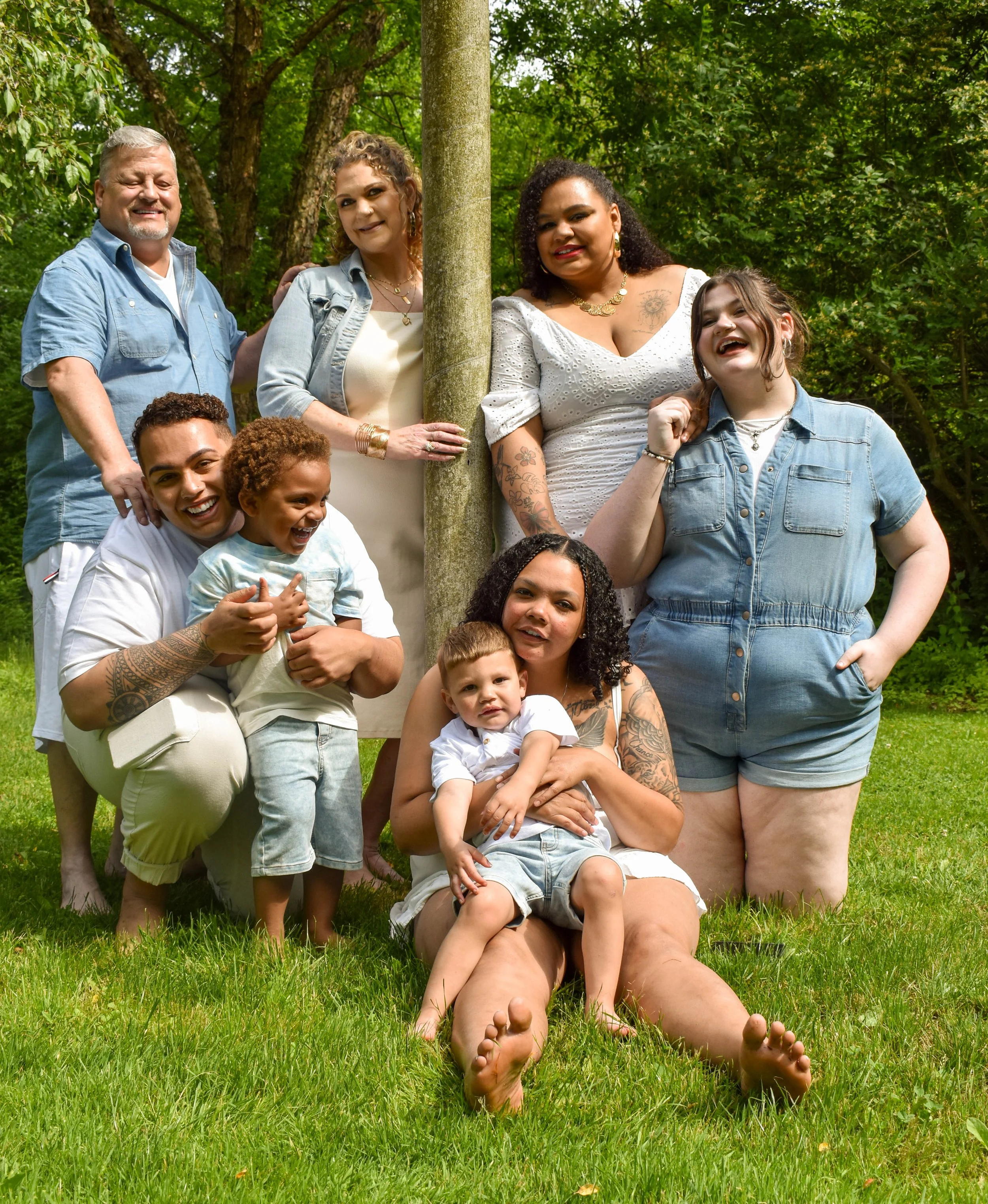 Group of diverse people and children sitting and standing outdoors on grass in a park with trees in the background.