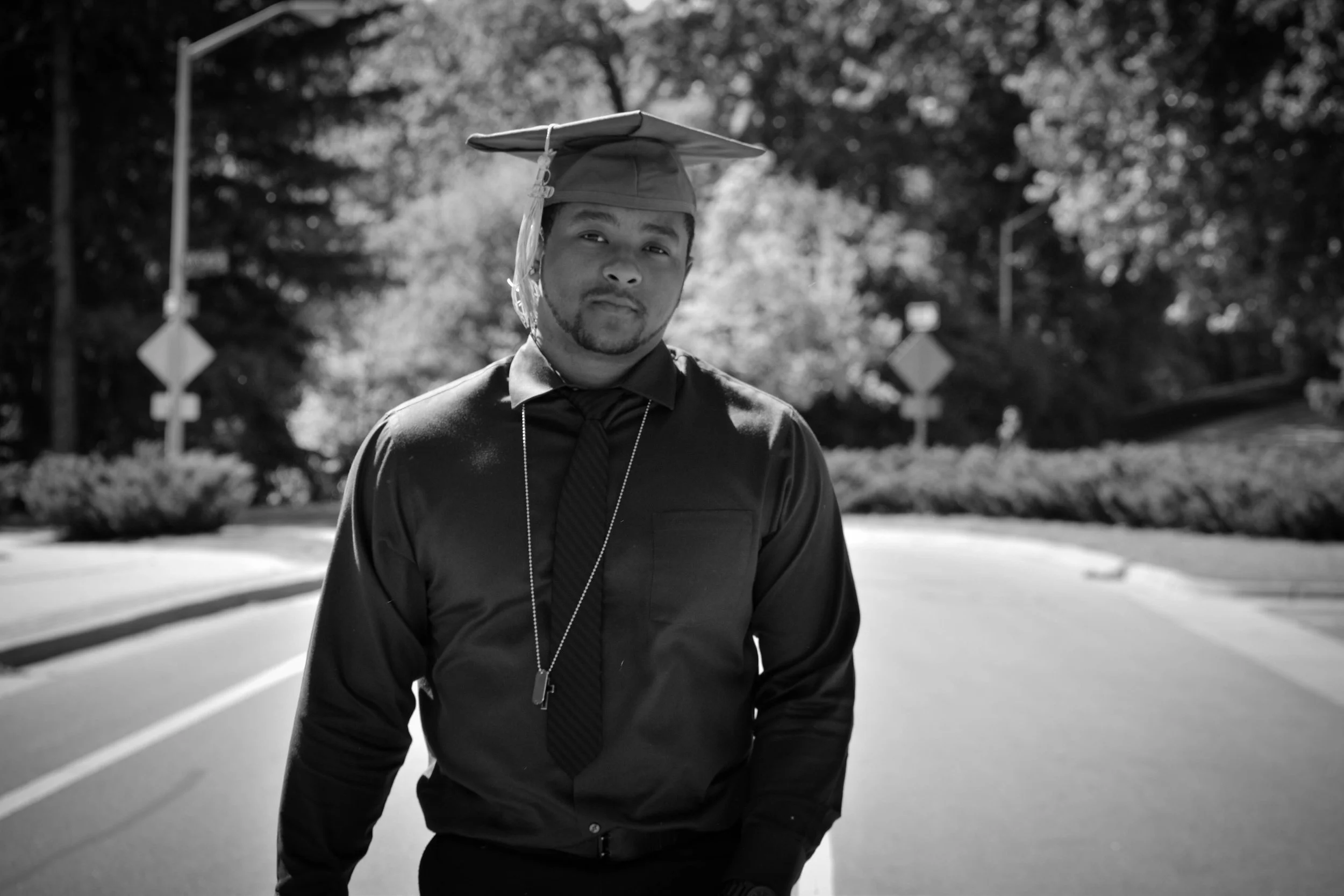A young man in a graduation cap and gown standing on a street with trees and street signs in the background, captured in black and white.