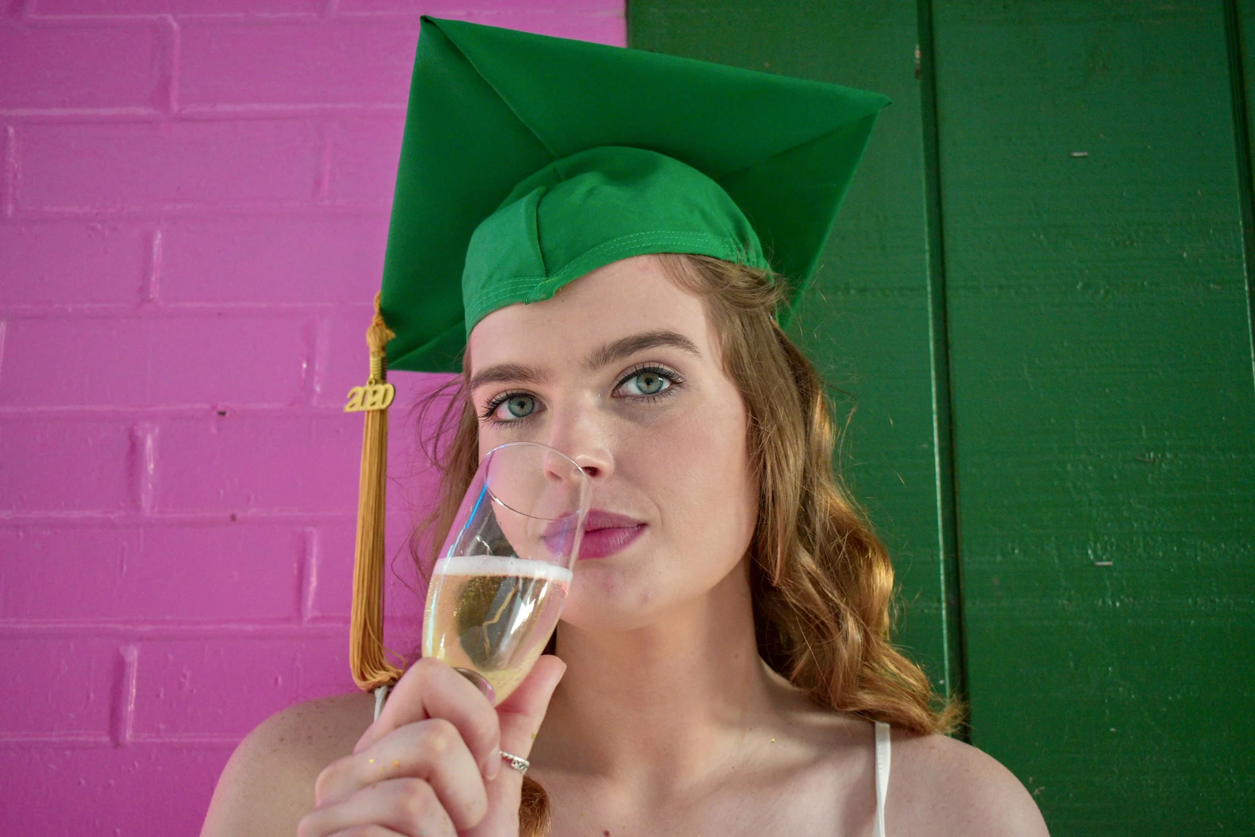 A young woman with red hair and blue eyes wearing a green graduation cap and holding a glass of champagne, standing in front of a pink and green wall.