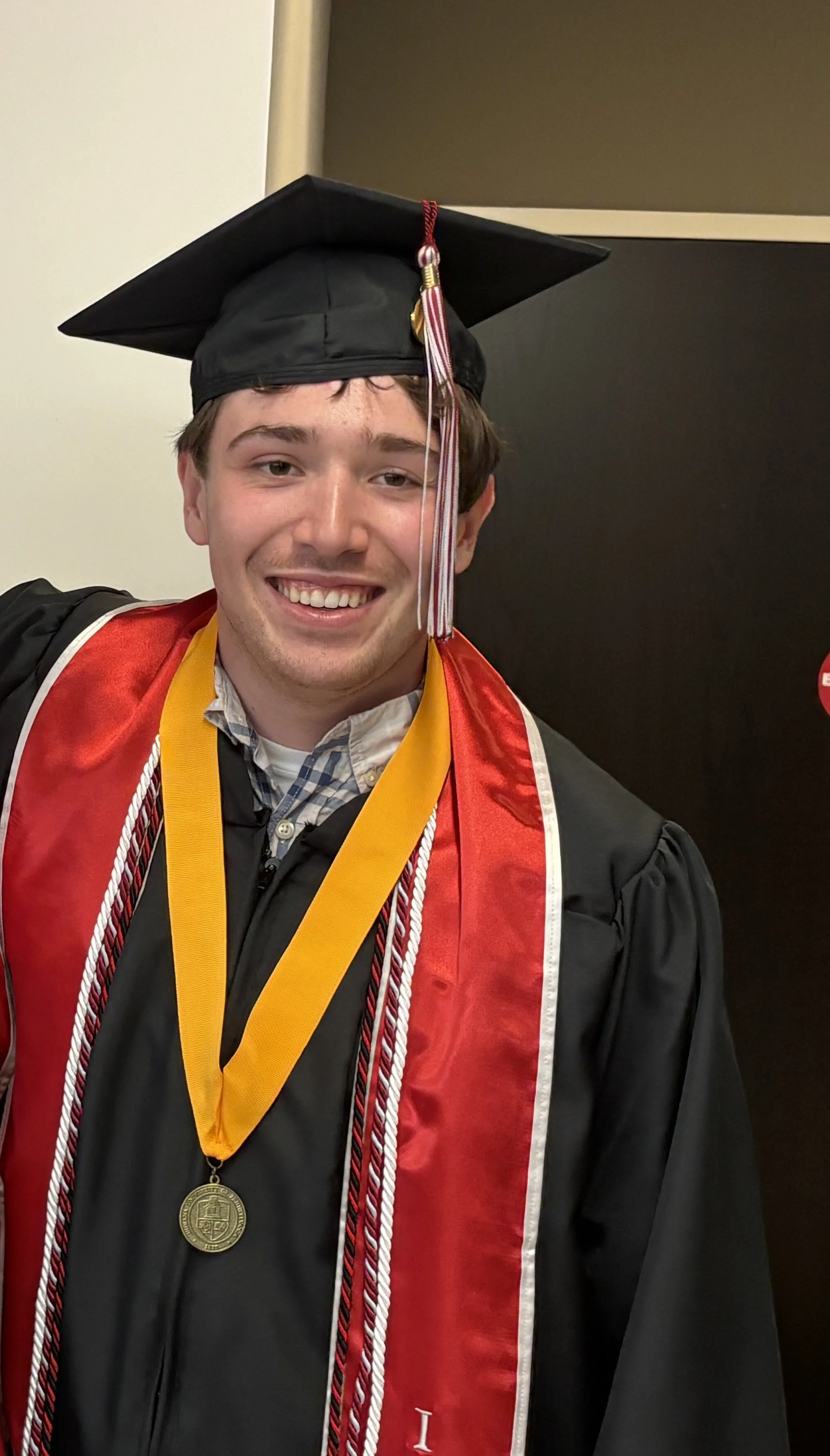 Young man in a graduation cap and gown with a red and black stole, wearing medals, smiling at the camera.