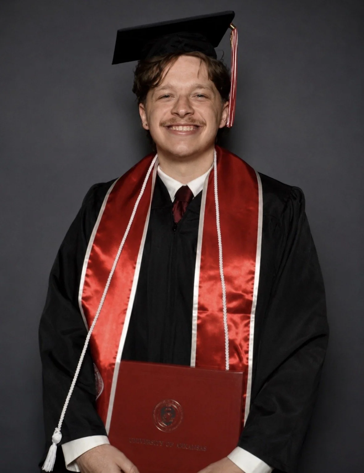A young man in a graduation gown and cap holding a red diploma folder with a university seal.