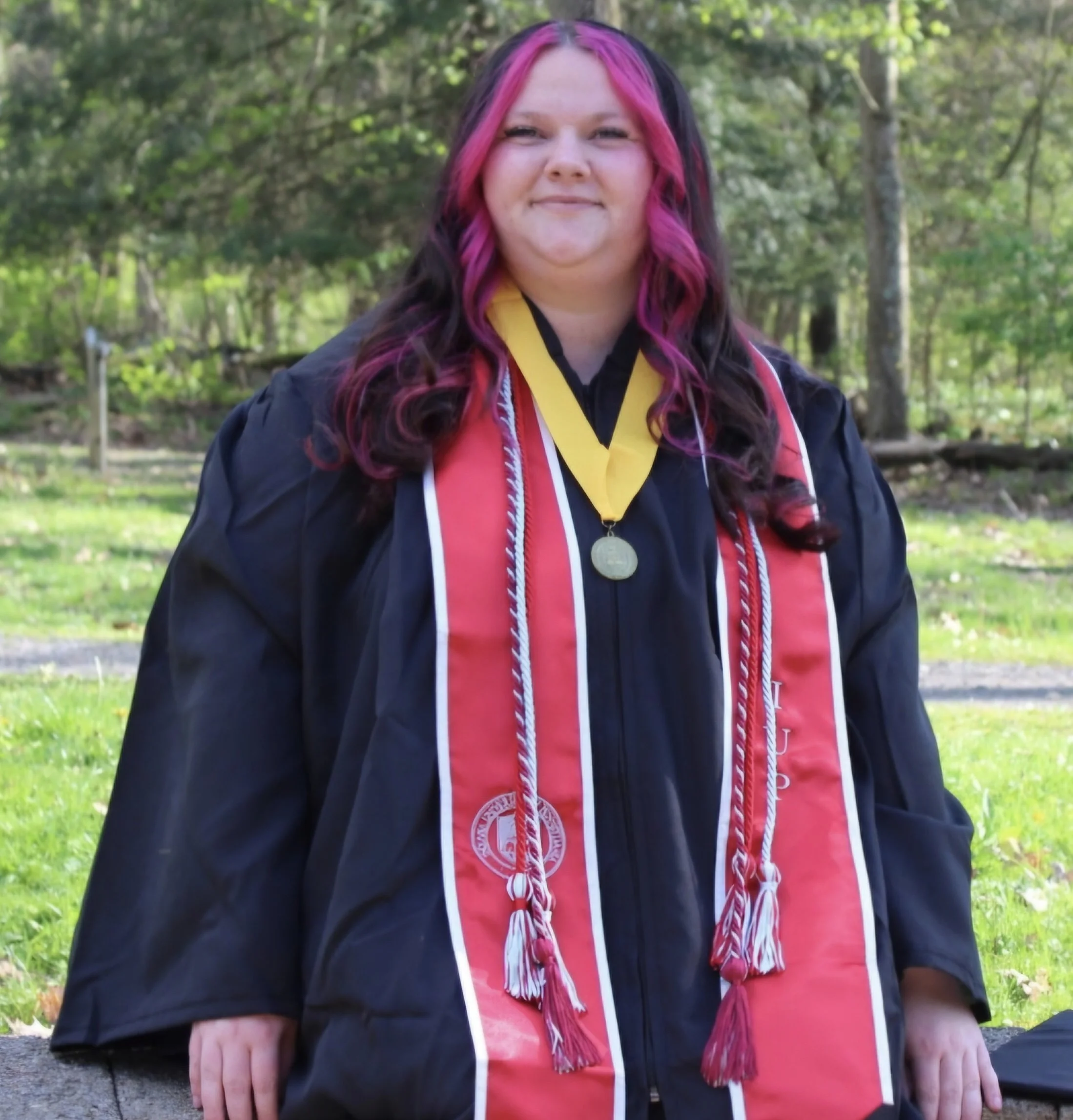 A woman with long, pink and black hair wearing a graduation gown and medals, sitting outdoors with trees and greenery in the background.