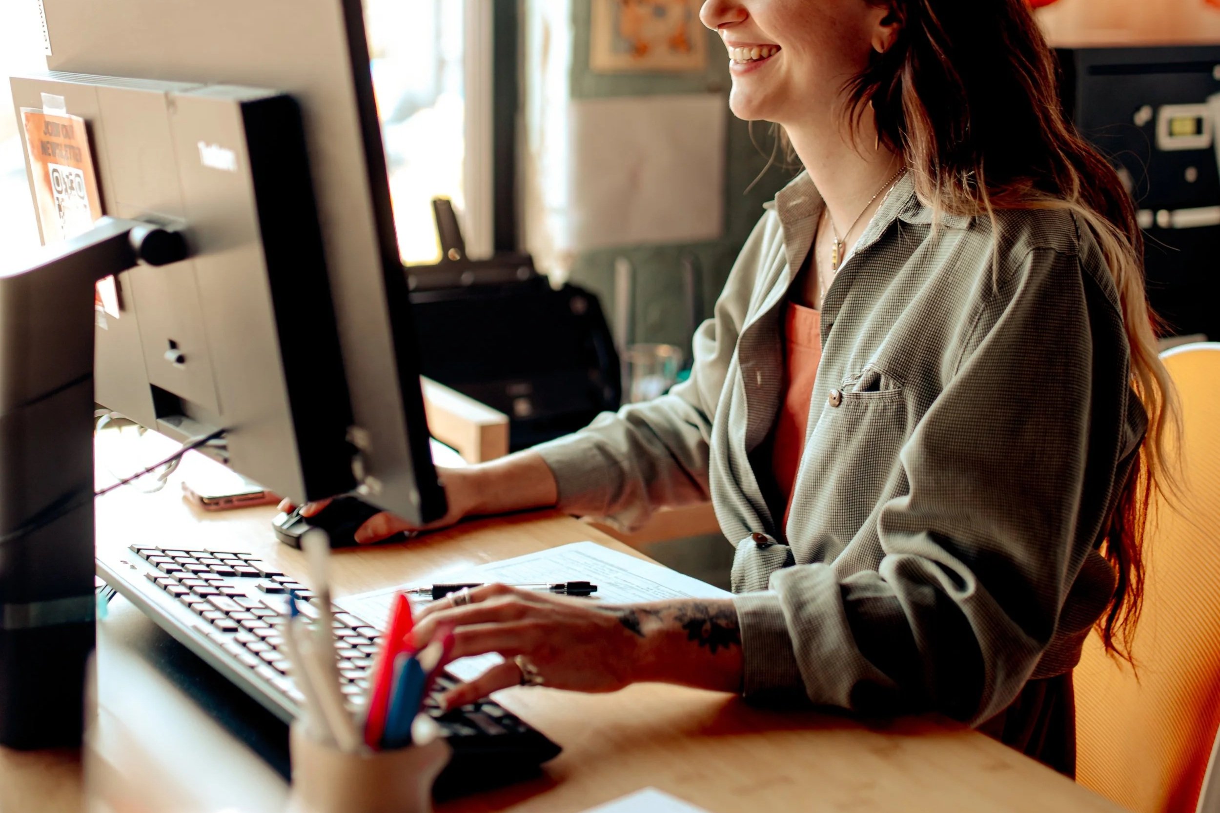 A woman with red hair smiling while working at a computer in an office or cafe setting, with papers, a keyboard, and writing supplies on the desk.