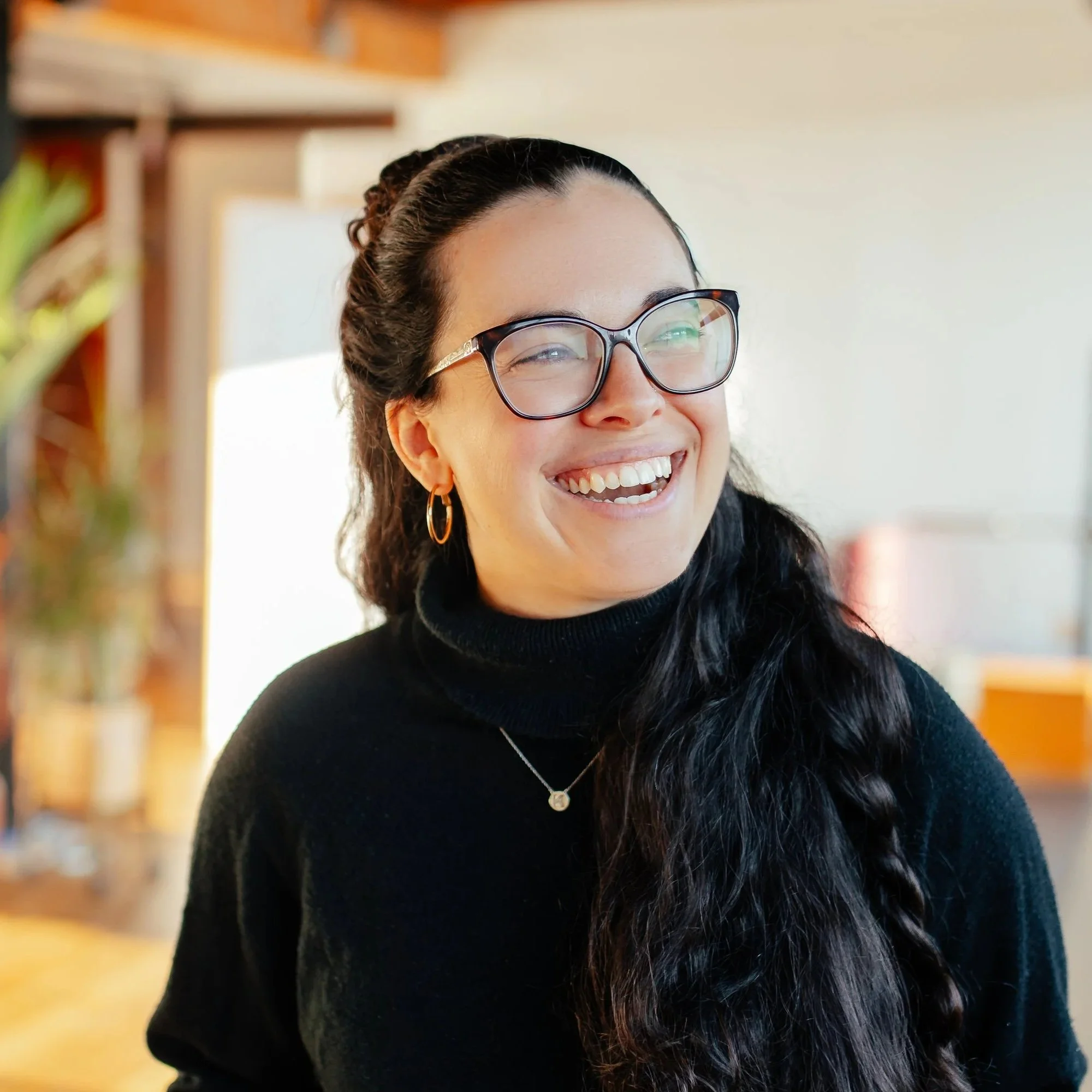 Close-up of a smiling woman with dark hair, glasses, hoop earrings, and a black top, indoors with a blurred background.
