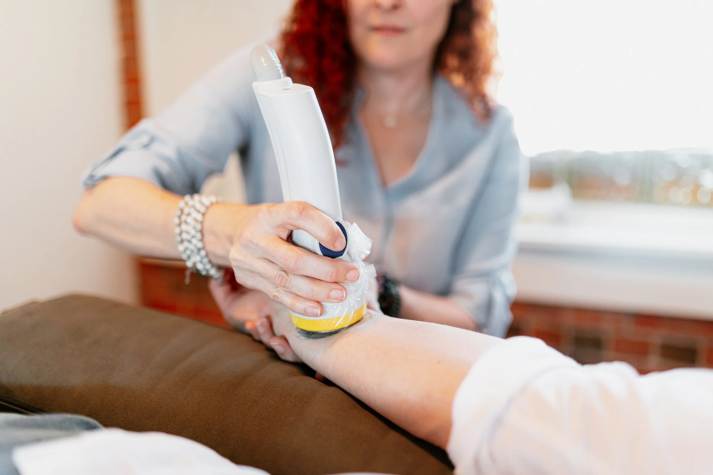 A woman with red hair wearing a light blue shirt, sitting at a table, performing a massage or medical treatment on someone's arm using a white device, in a bright room.