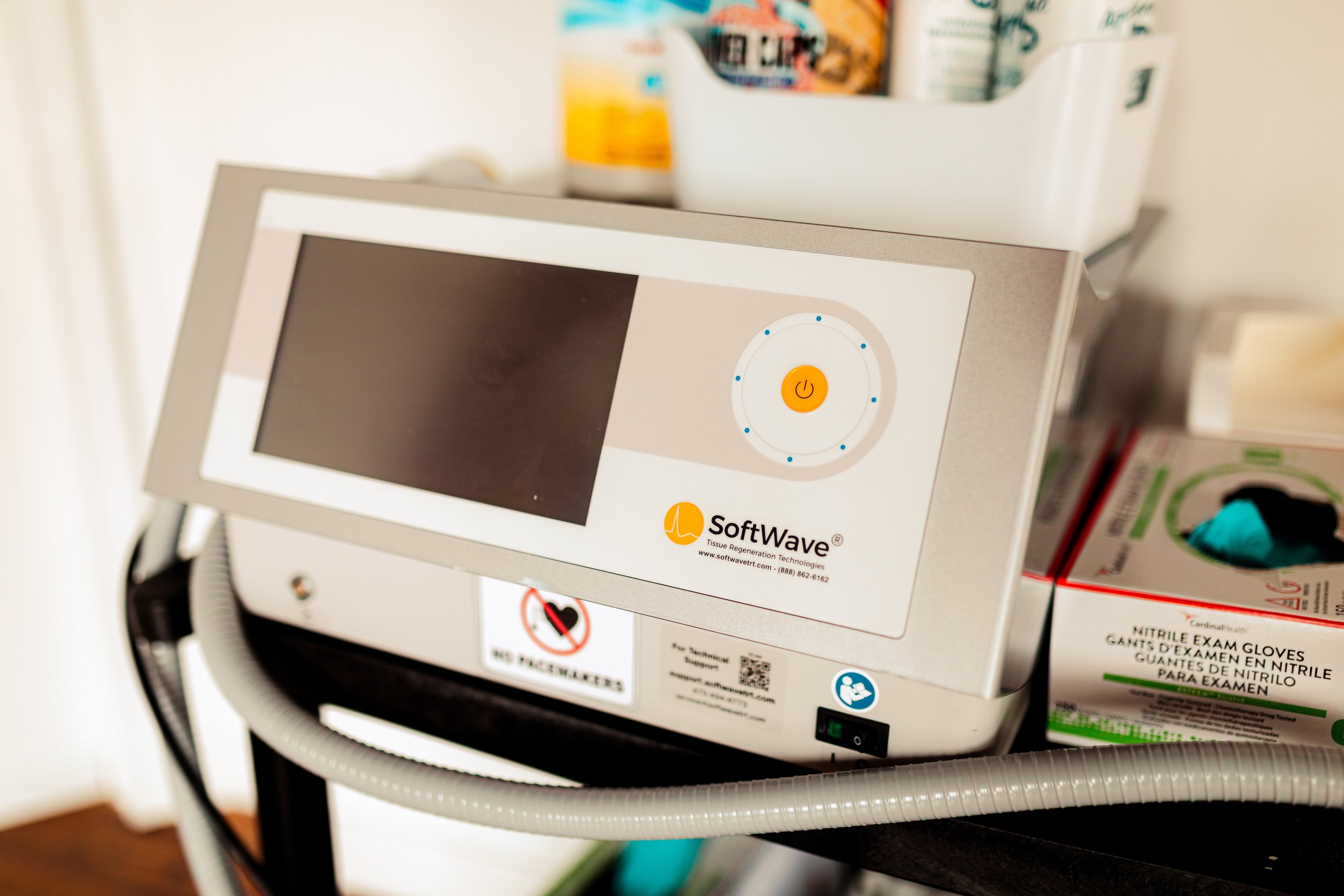 Close-up of a SoftWave Tissue Regeneration Technologies device with a touchscreen and orange power button, placed on a black cart with shelves, surrounded by medical supplies and boxes.