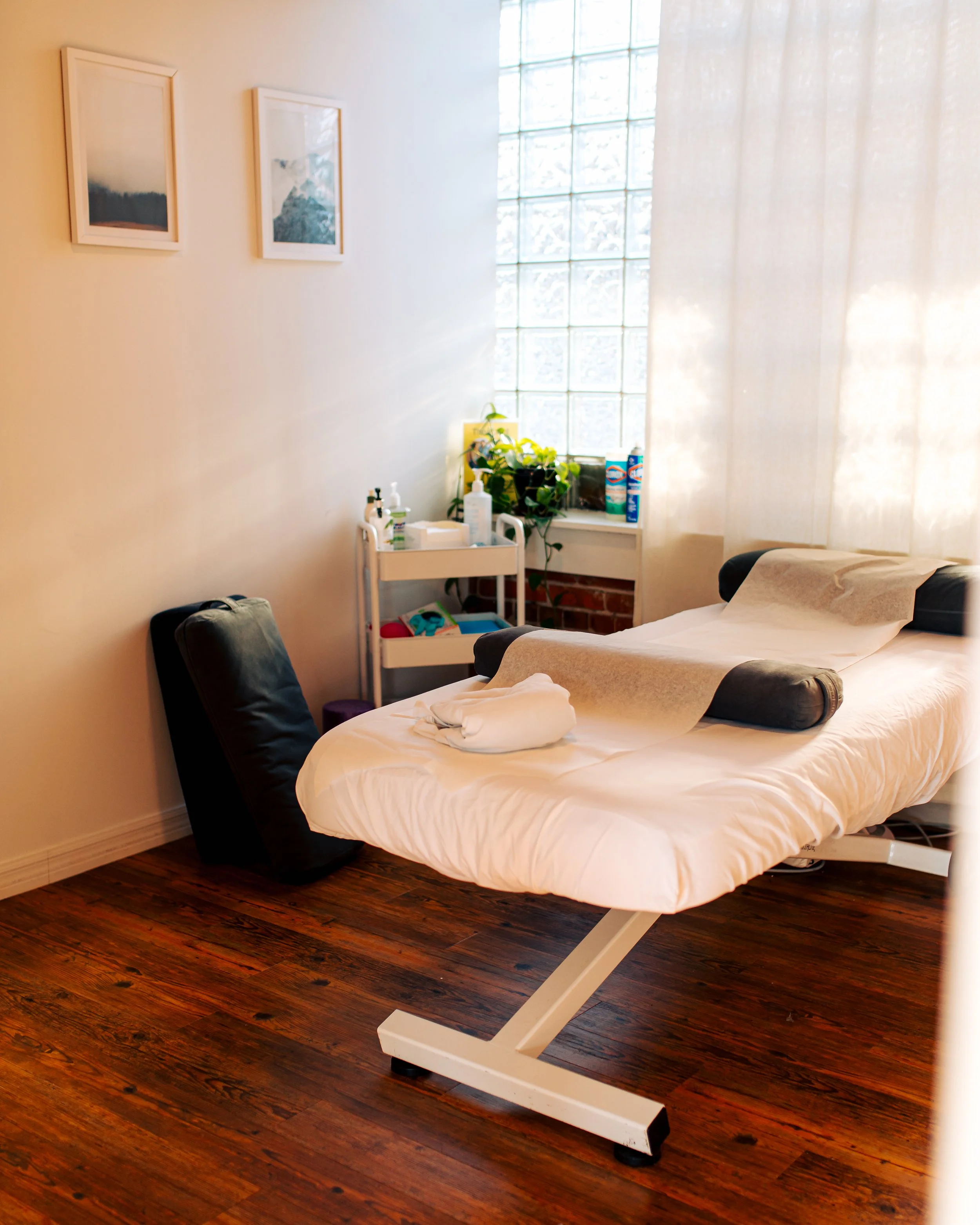 A hospital bed with white sheets and a pillow, positioned in a room with wooden floors, a window with glass blocks, and a white curtain. Nearby is a white bedside table with medical supplies and a green potted plant.