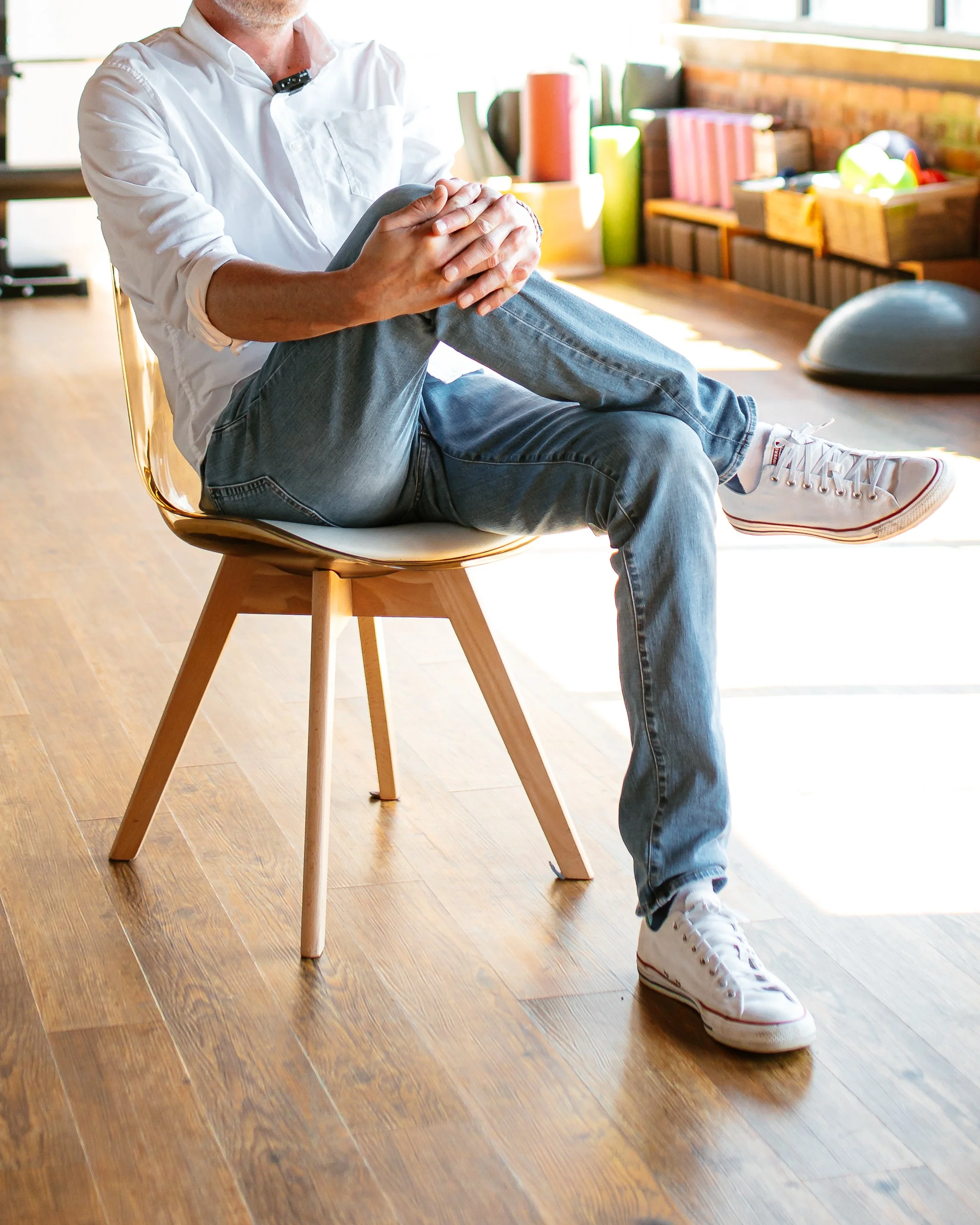 Person with light skin wearing white sneakers and jeans, sitting on a wooden chair with a rounded seat, stretching their leg on their lap, in a brightly lit room with wooden flooring and a background of storage boxes and books.