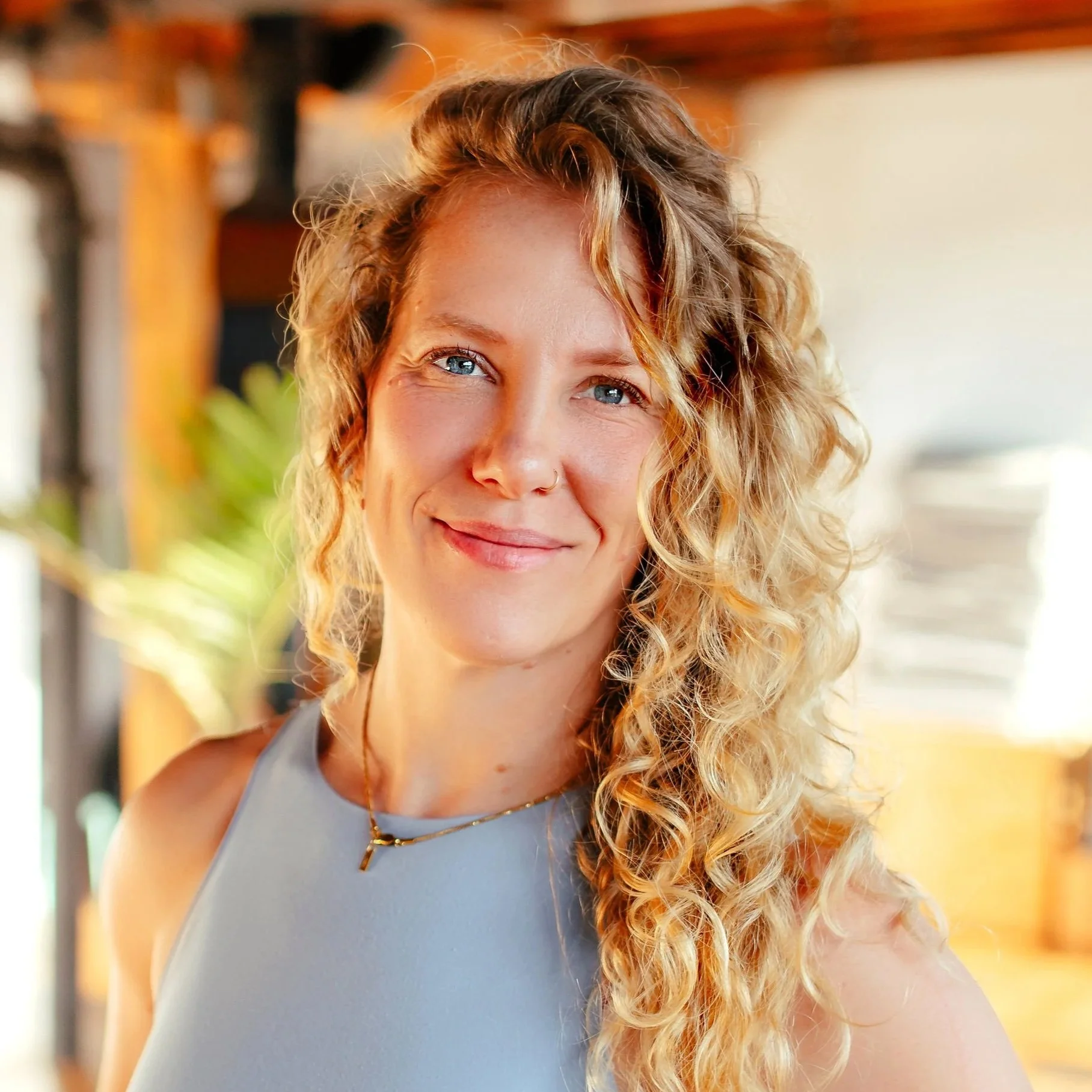 Close-up of a smiling woman with curly blonde hair, blue eyes, wearing a gray sleeveless top, a gold necklace, and a gold pendant, in a well-lit indoor space.
