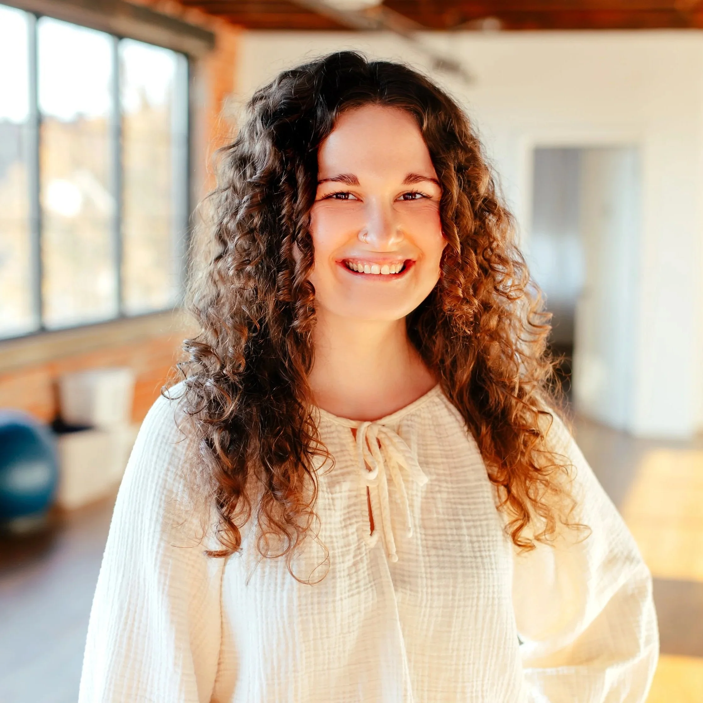 Close-up of a woman with curly hair smiling indoors near windows.