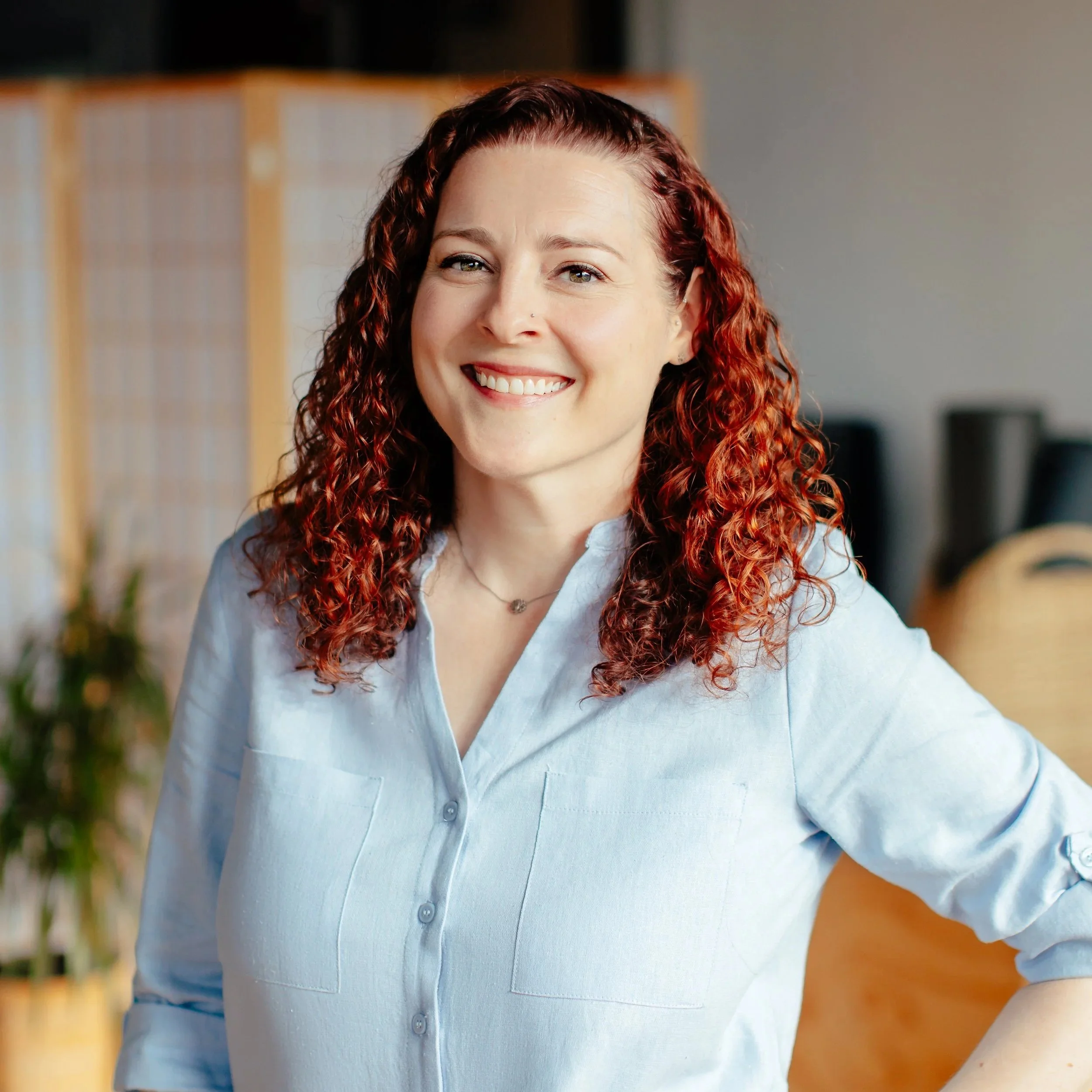 Close-up of a smiling woman with curly red hair, wearing a light blue button-up shirt, in an indoor setting.