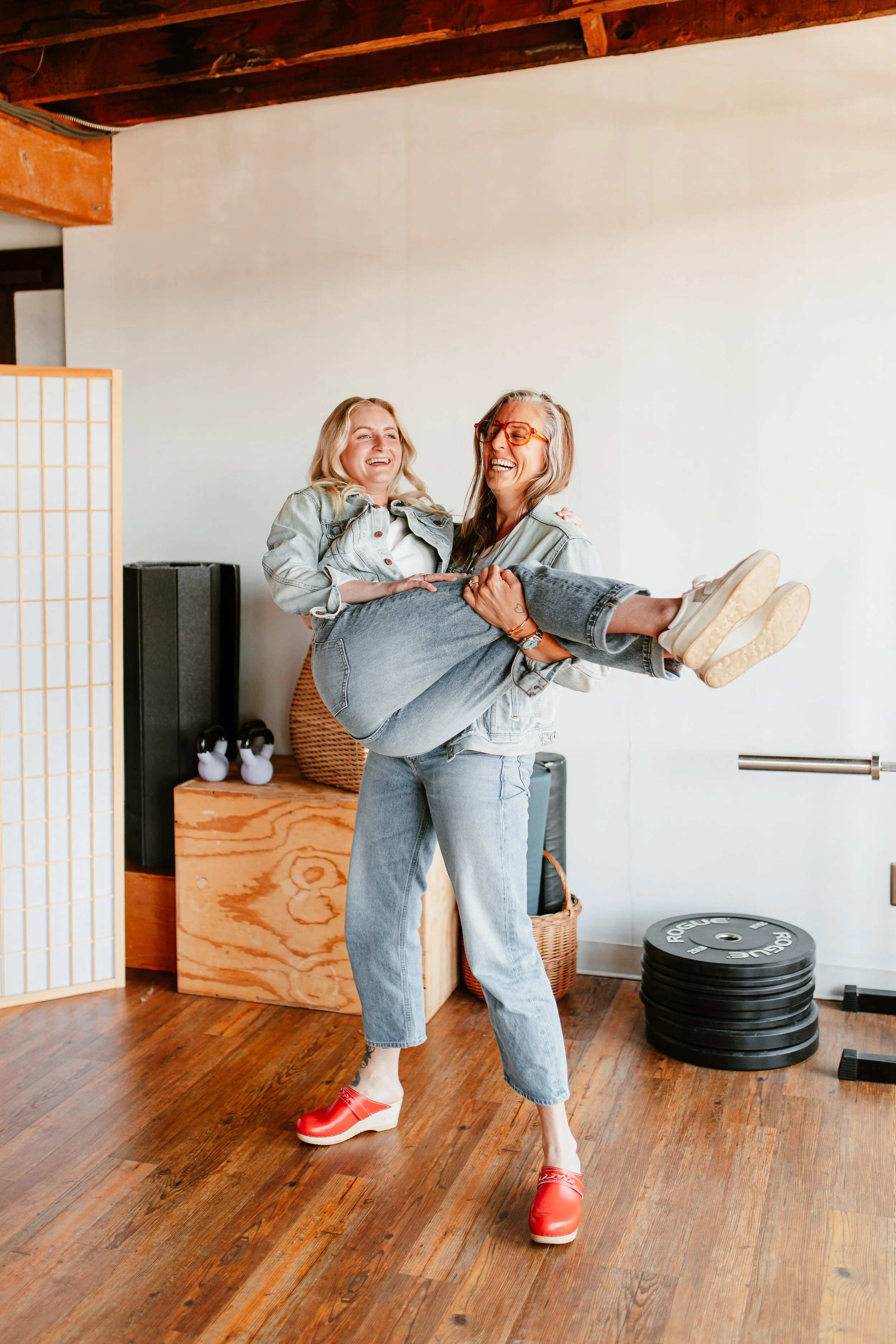 Two women laughing and smiling indoors; one is carrying the other, who is wearing white sneakers and red platform shoes, against a background with wooden flooring, a black speaker, stacking weights, and a wicker basket.