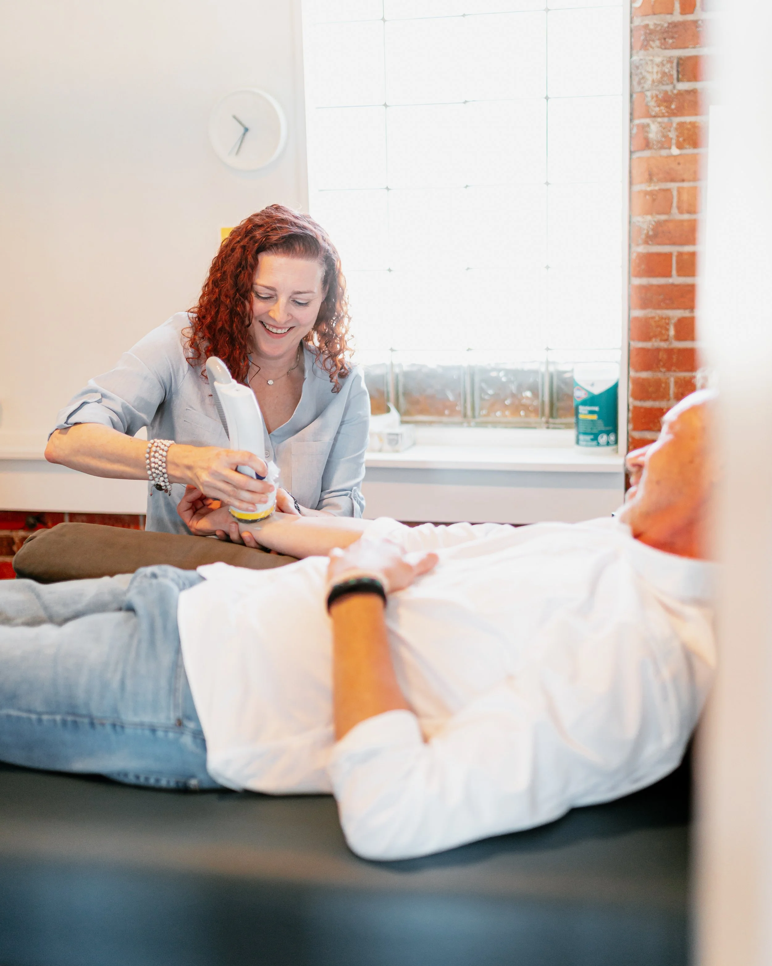 A woman with red curly hair smiling while giving a manicure to a person lying on a bed. The bed has white and orange sheets. There is a large window with bright natural light and a brick wall on the right.