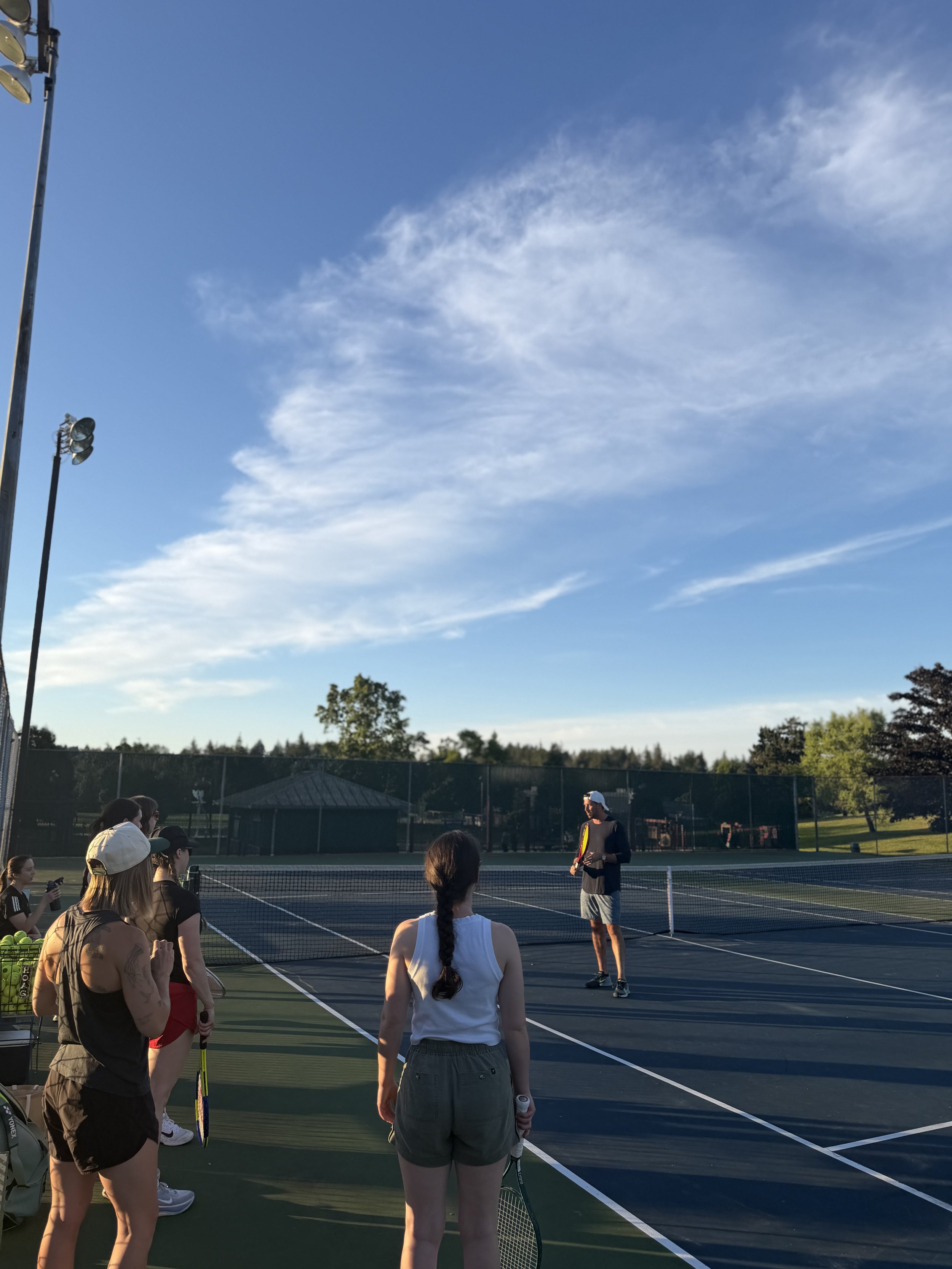 Group of people gathered on a tennis court, listening to a man in a black shirt and gray shorts holding a tennis racket, with tennis balls and paddles nearby. The scene is outdoors on a clear day with blue skies and some clouds.