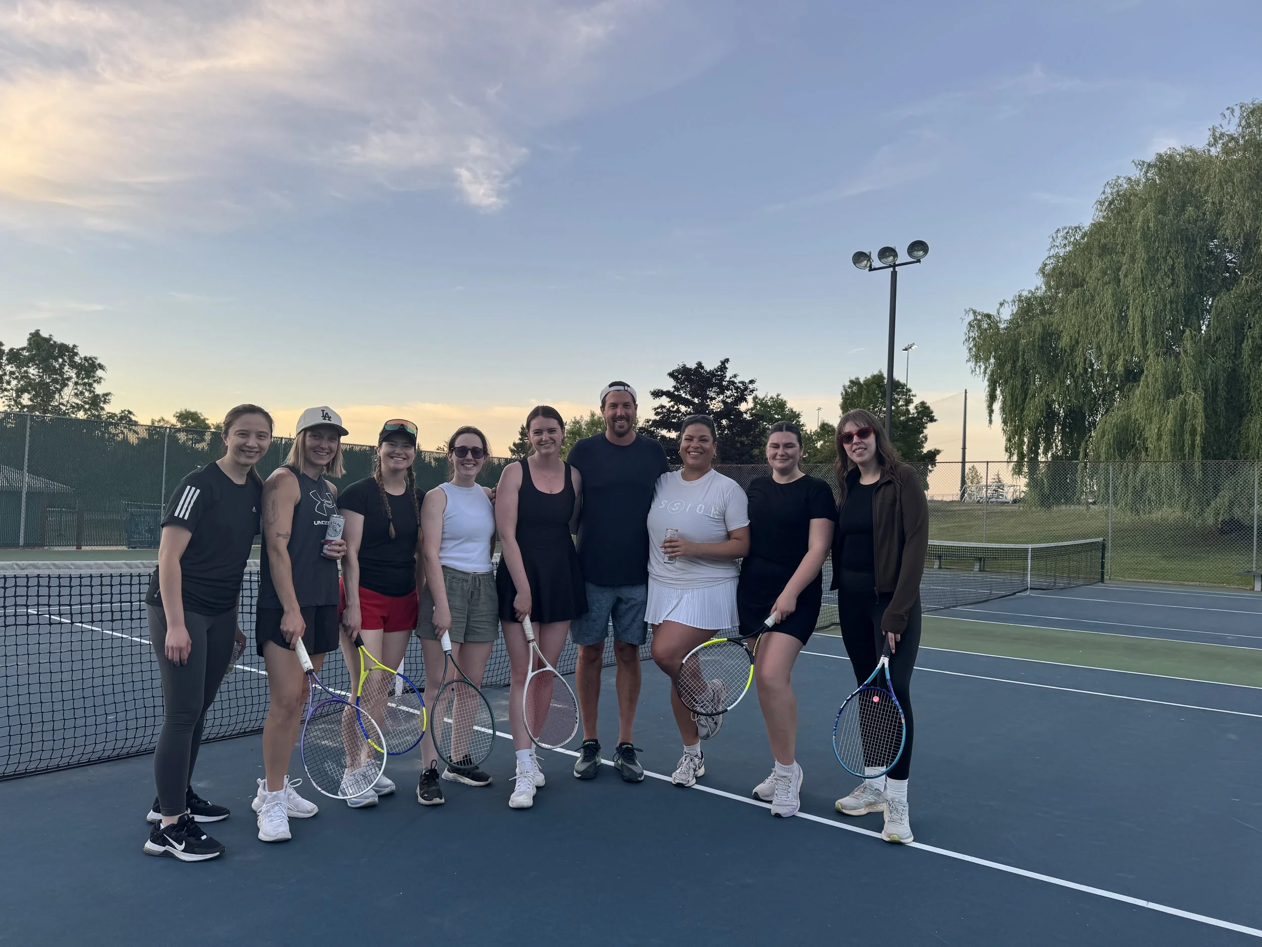 Group of people on a tennis court at sunset holding tennis rackets, smiling, dressed in athletic clothing, with trees and a blue sky in the background.