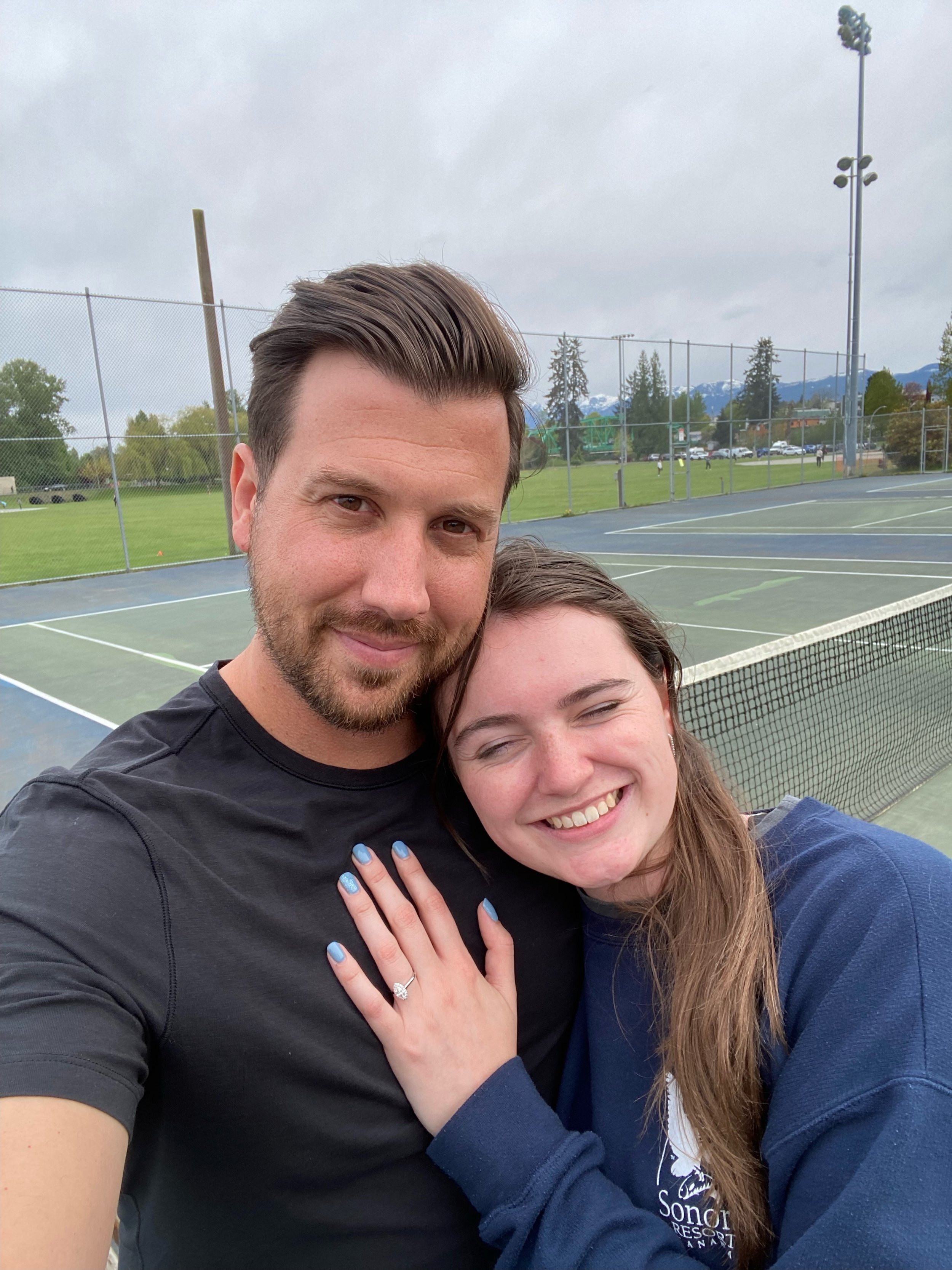 A man and woman smiling and embracing each other at a tennis court, with cloudy skies and mountain scenery in the background.