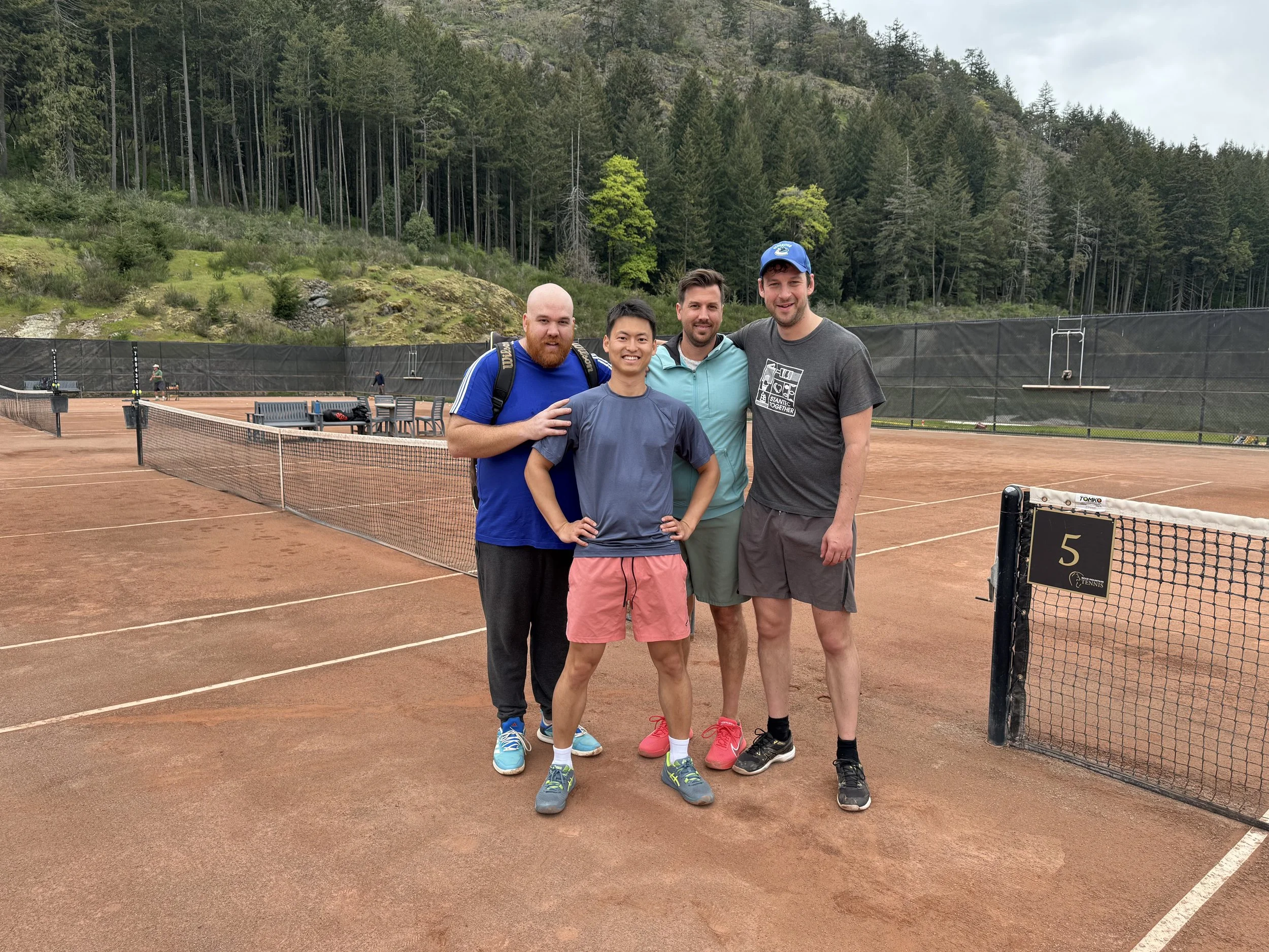 Four people standing on a clay tennis court with a forested hillside in the background. They are smiling, with arms around each other, dressed in athletic clothing suitable for tennis.