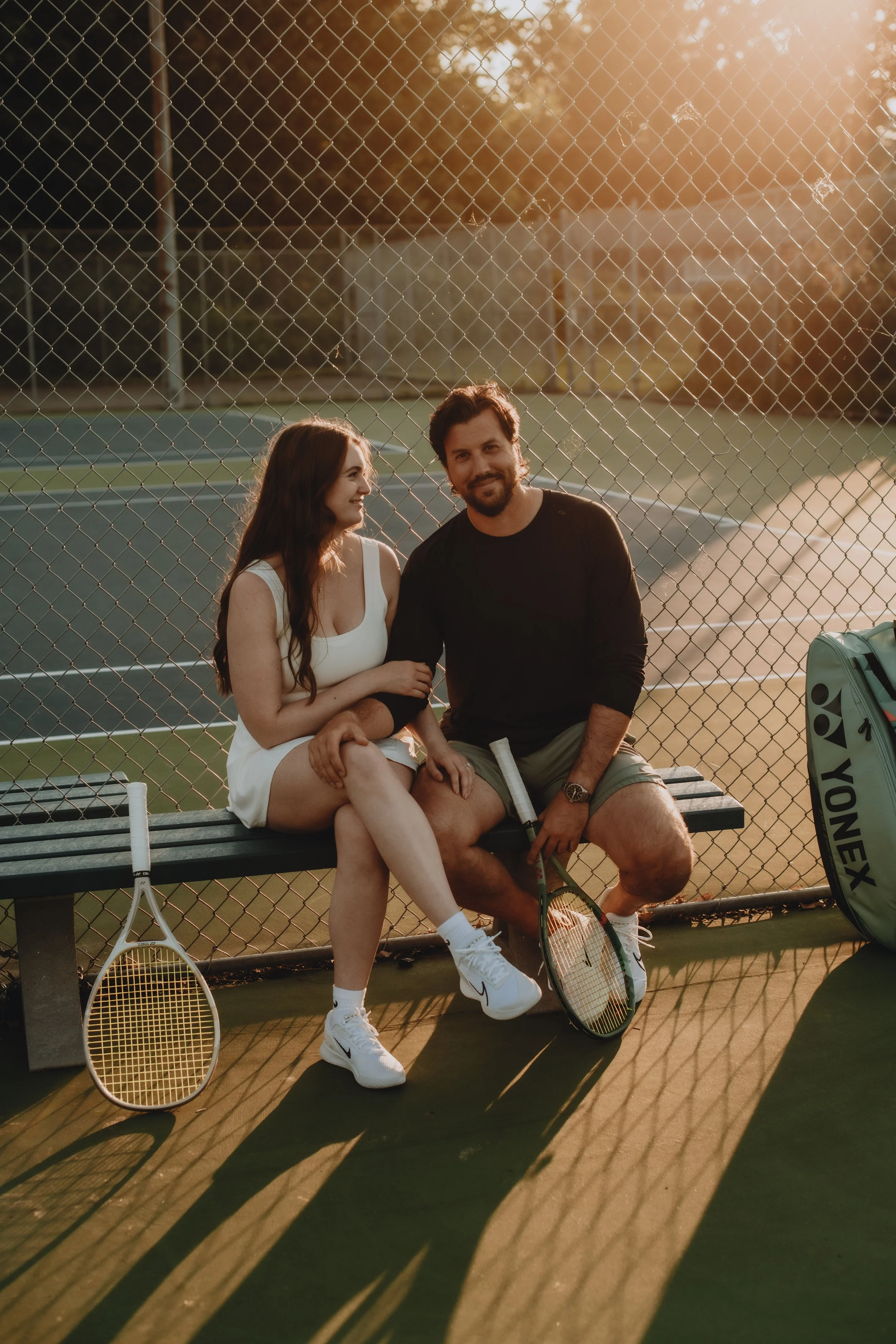A man and a woman sitting on a tennis bench behind a chain-link fence at sunset, with tennis rackets beside them on the ground.