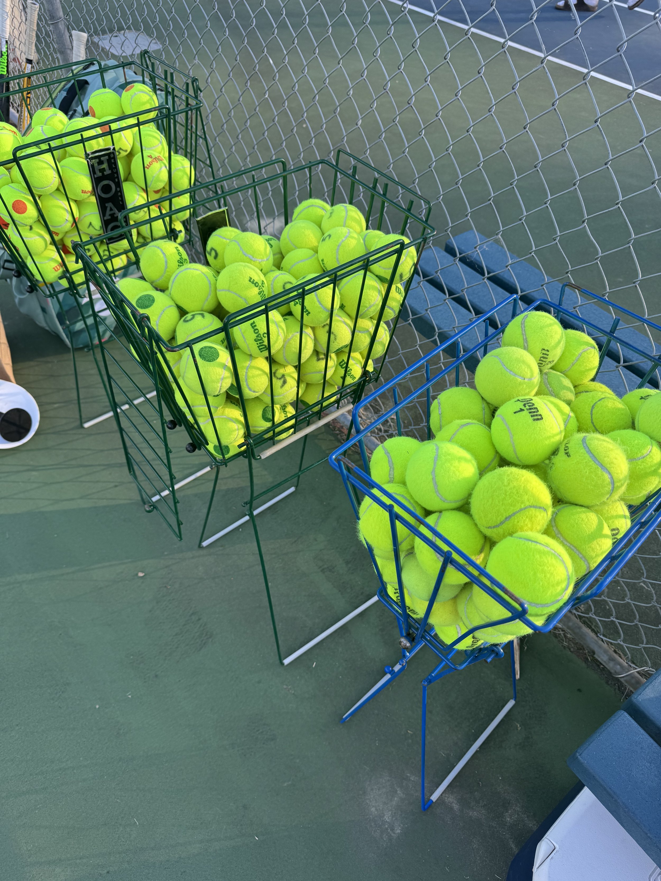 Two baskets of bright yellow tennis balls on a tennis court, with a chain-link fence in the background.