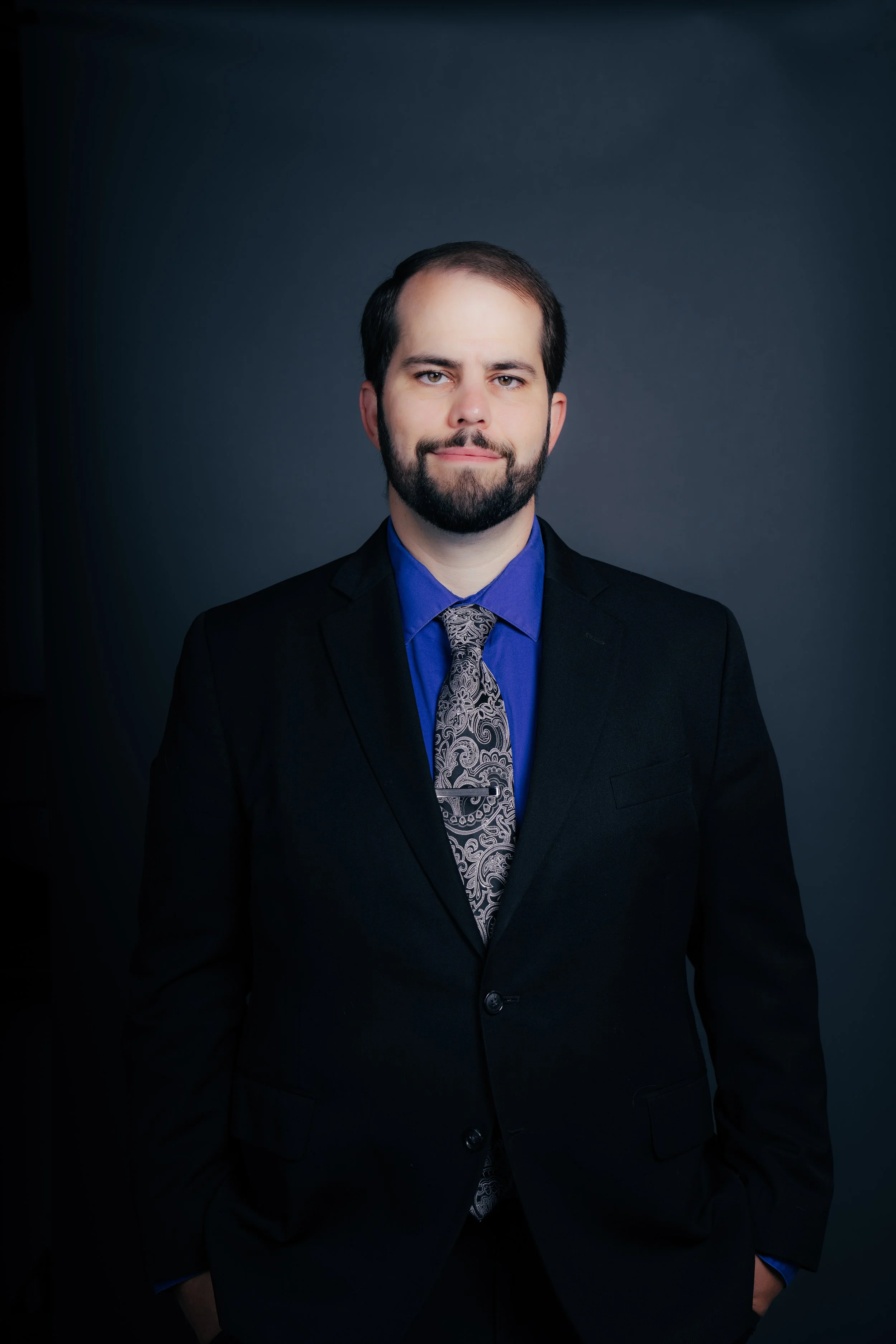 Portrait of a man with dark hair and beard, wearing a black suit, blue shirt, and patterned tie, standing against a dark grey background.