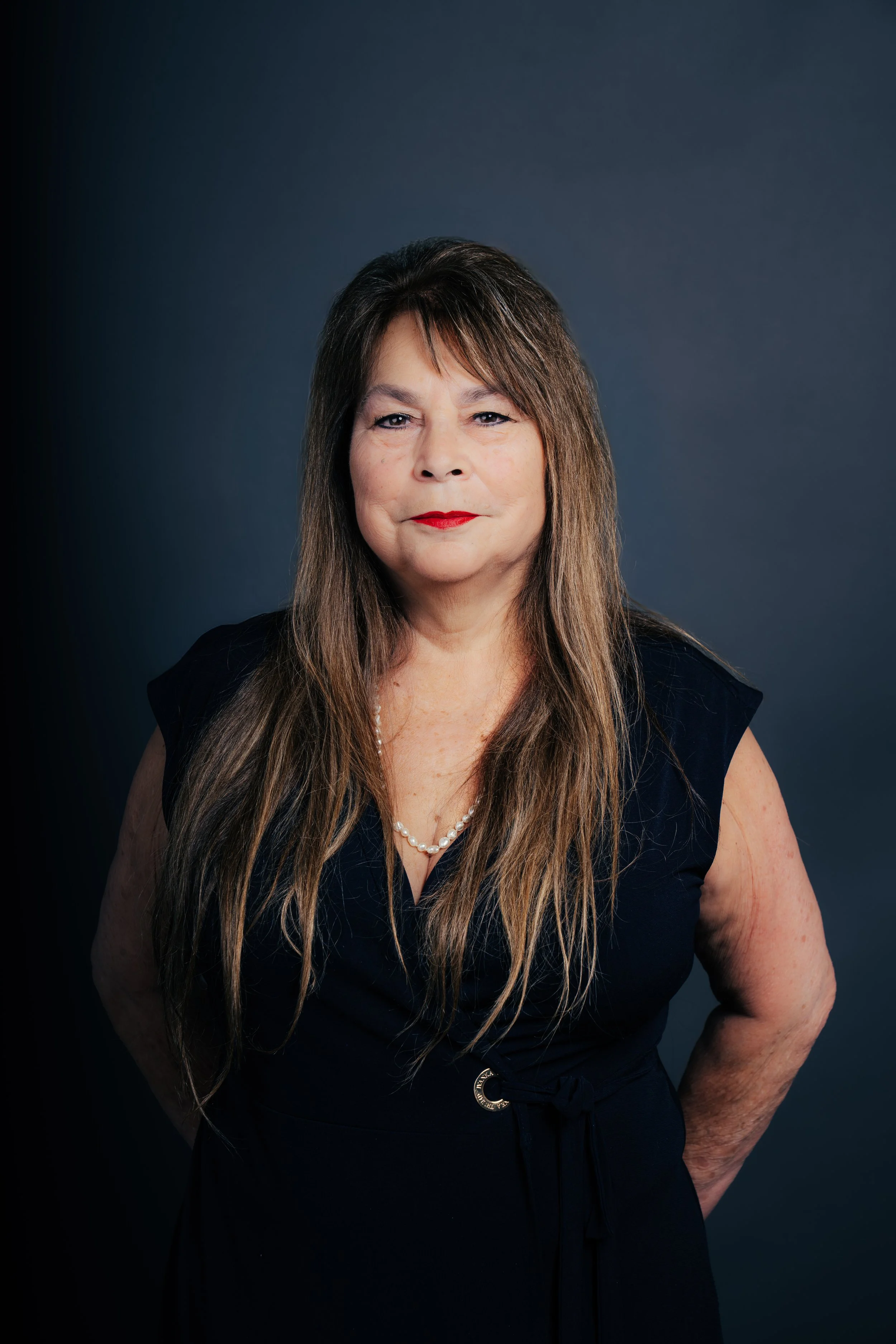 Portrait of a middle-aged woman with long brown hair, wearing a black dress and a pearl necklace, standing against a dark background.