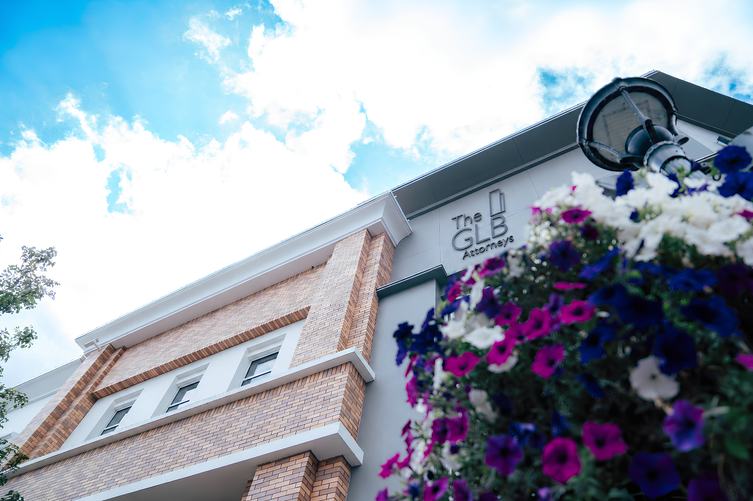Low-angle view of a modern brick and white building with a sign that reads 'The GLB Attorneys,' with a bright cloudy sky overhead and colorful flowers in the foreground.