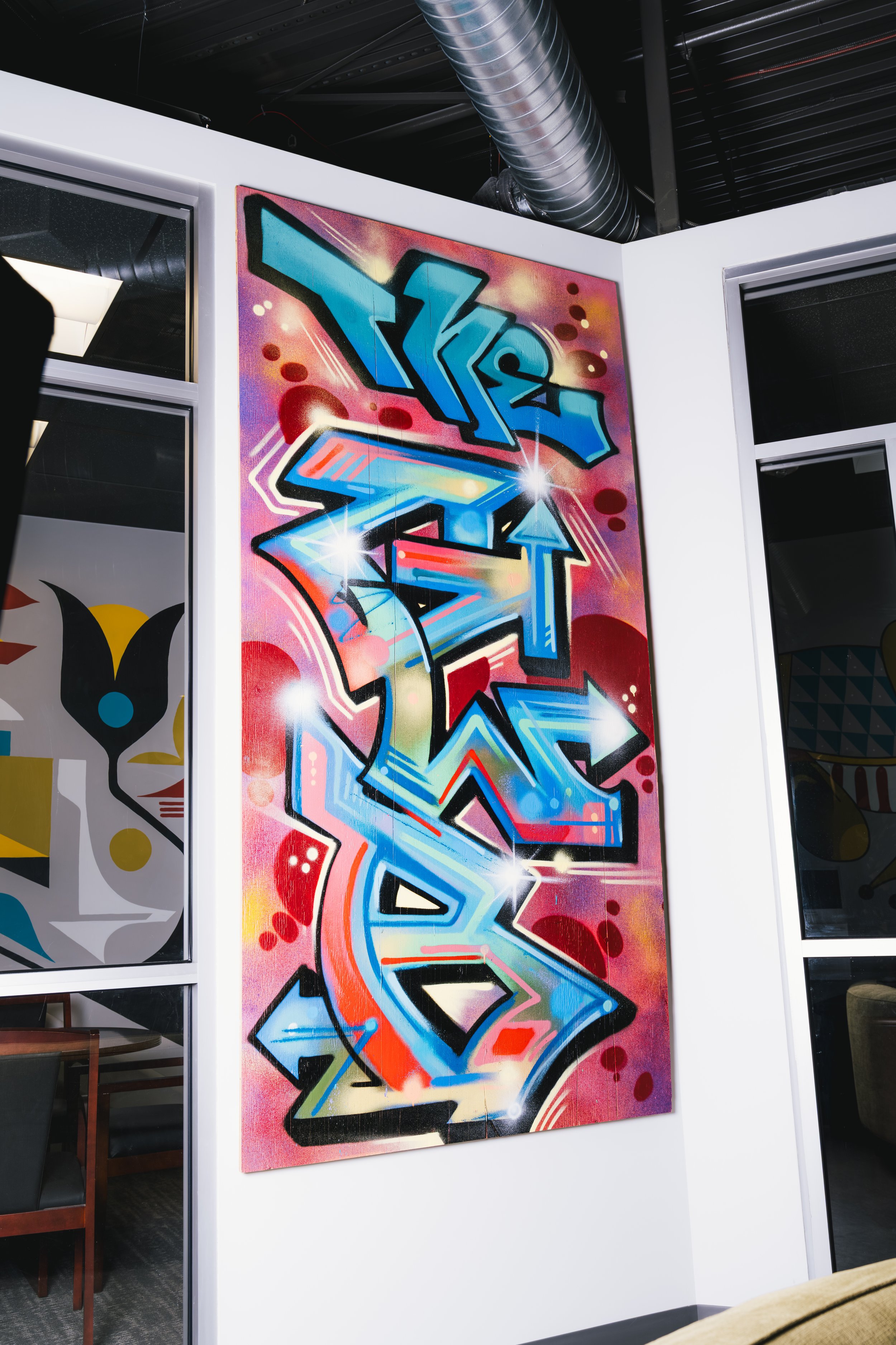 Colorful graffiti art mural with abstract shapes and arrows, vibrant pink, blue, red, and yellow hues on a wall in an indoor setting.