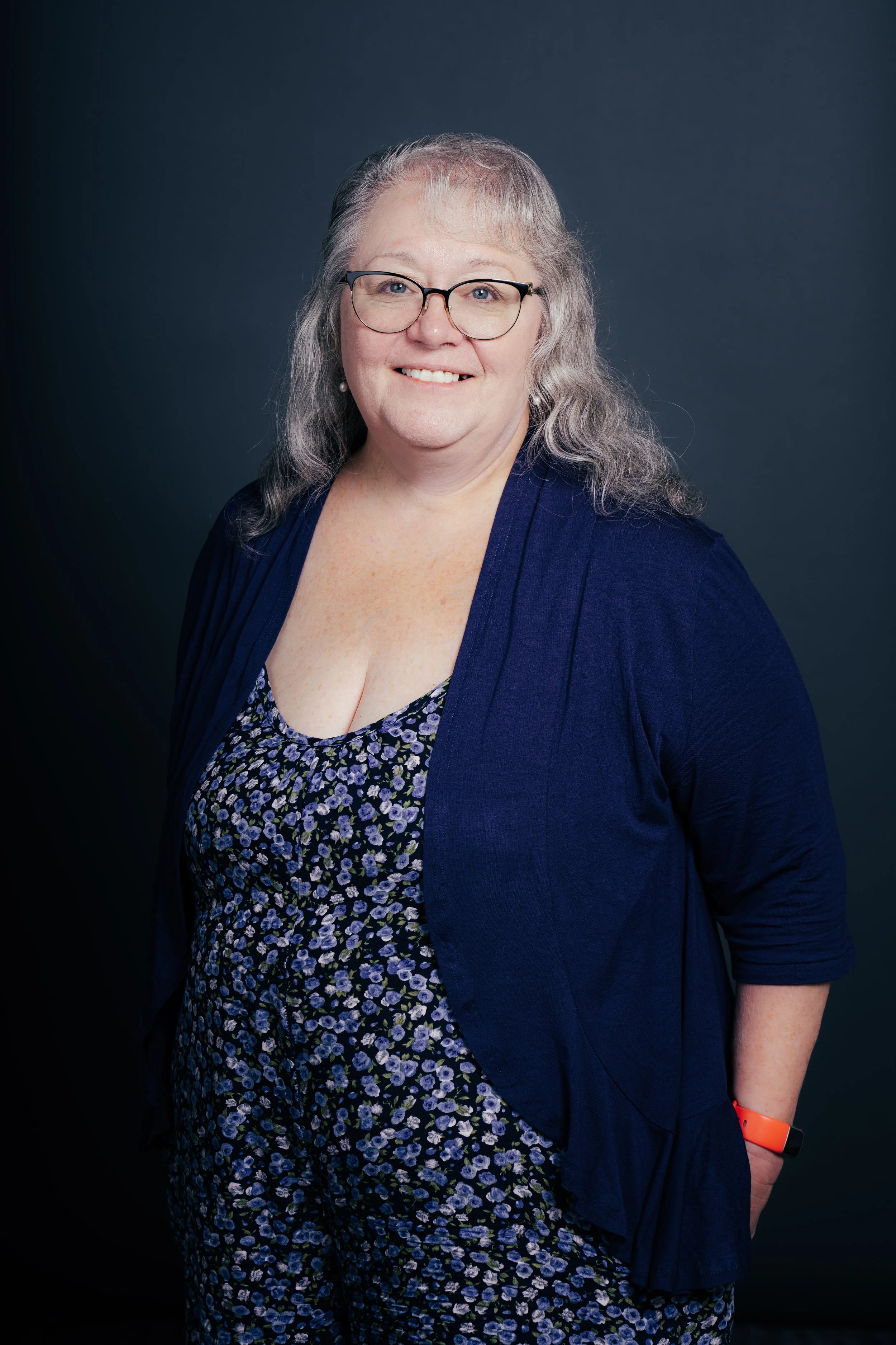 Portrait of a woman with gray hair and glasses, smiling, wearing a floral dress and a blue cardigan, against a dark background.