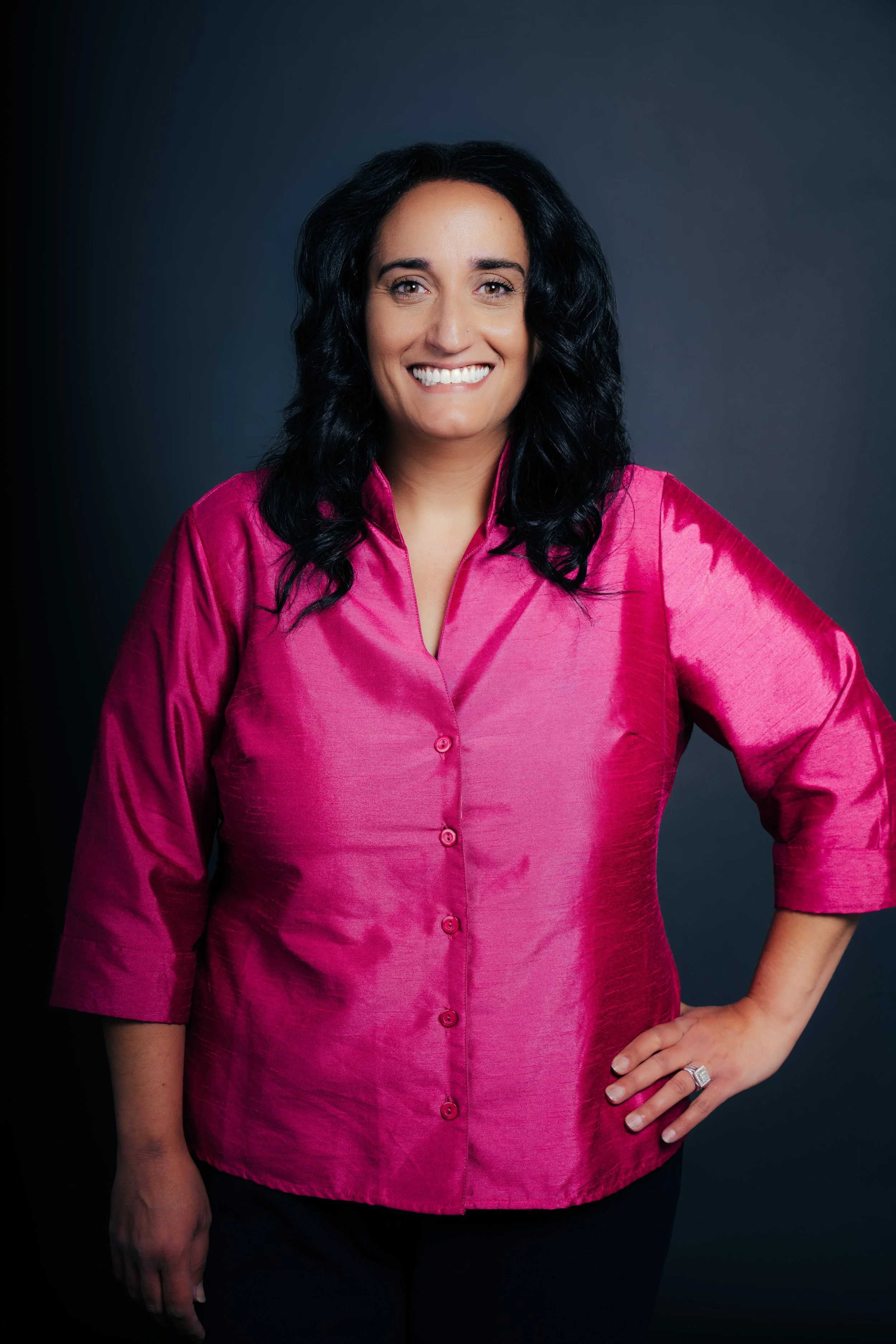 A woman wearing a bright pink silk shirt standing with one hand on her hip against a dark background.