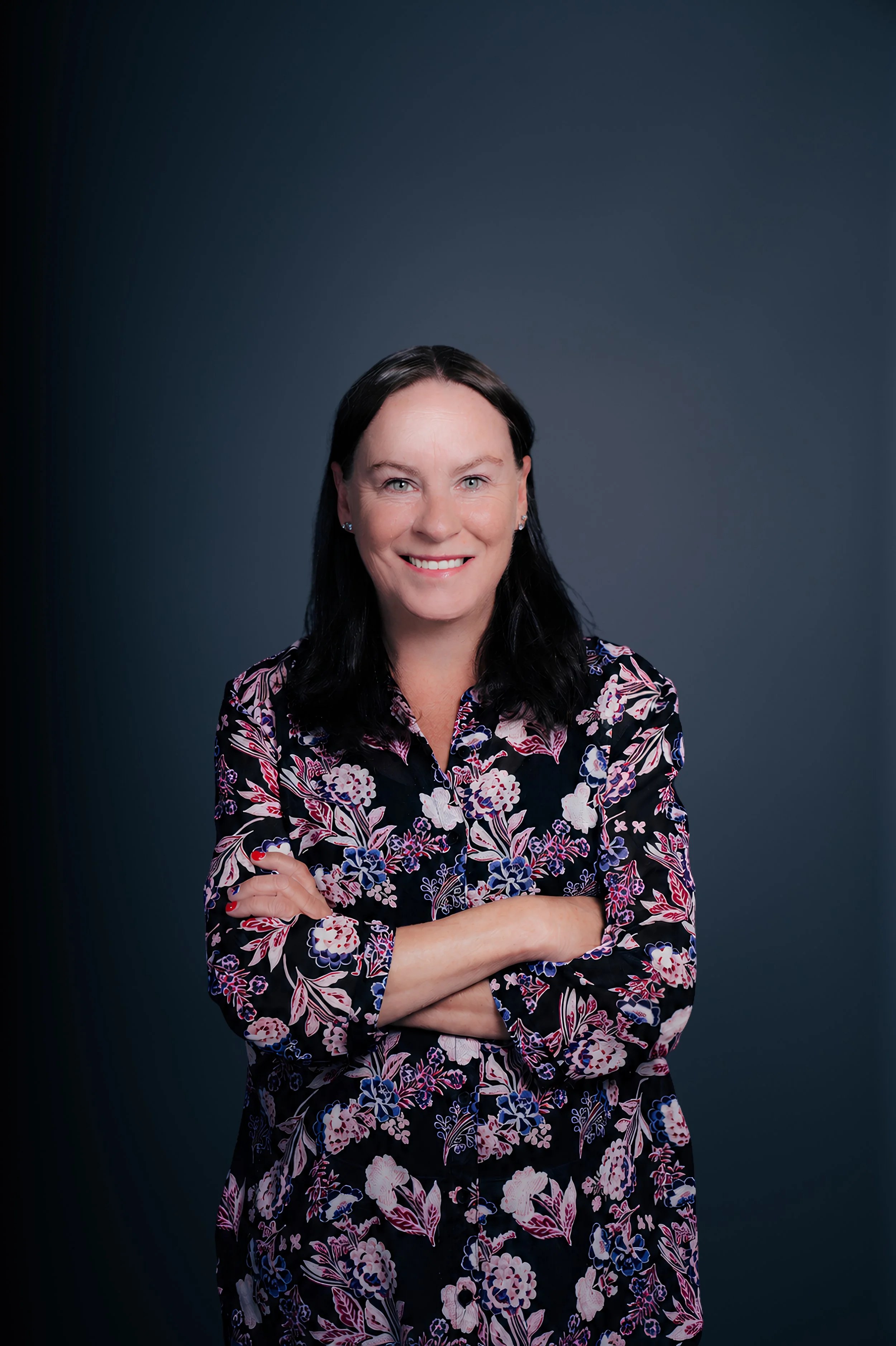 Portrait of a woman with dark hair and light skin, wearing a floral blouse, smiling, with arms crossed, against a dark gray background.