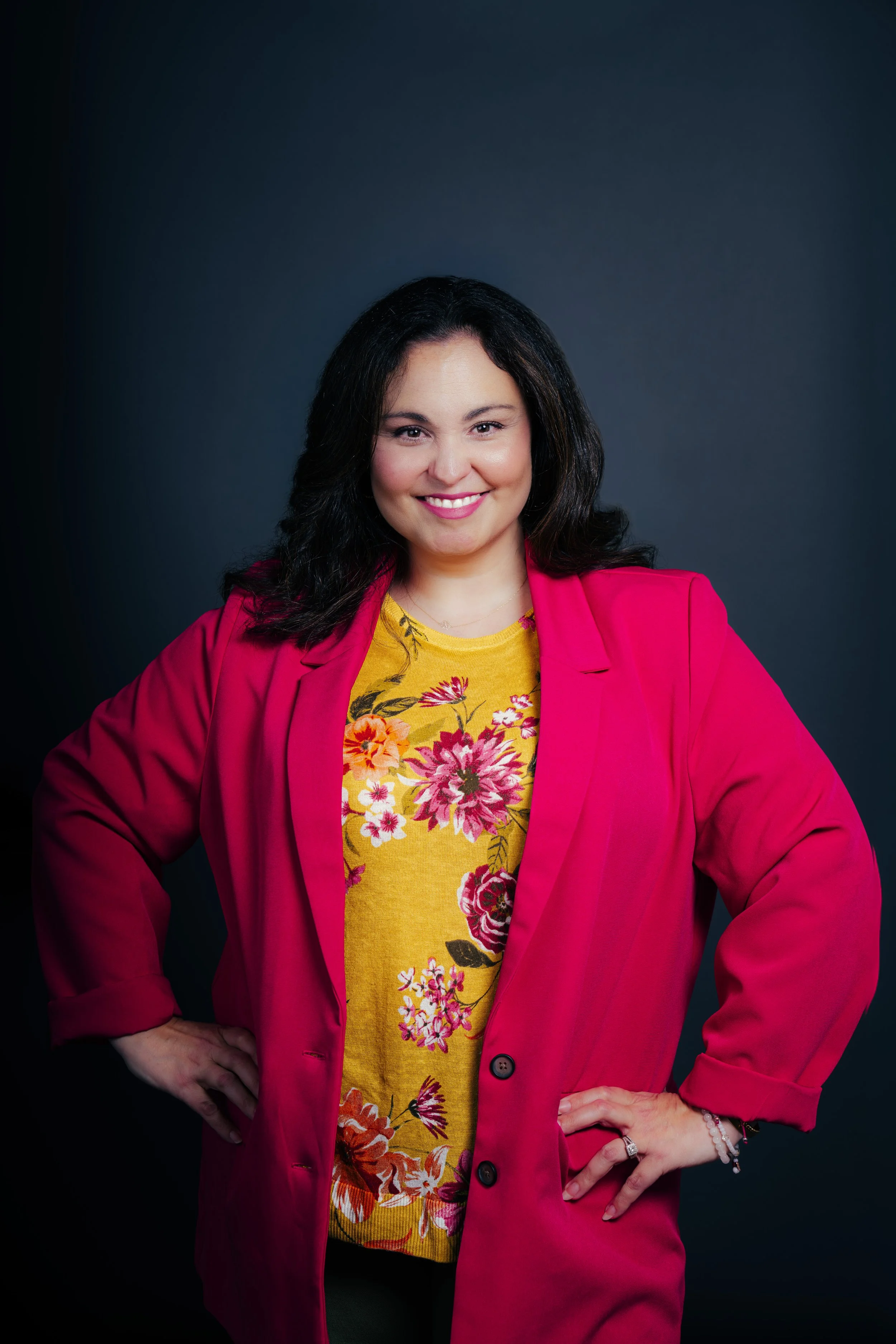 A woman with dark hair and light skin wearing a bright pink blazer over a yellow floral top, standing with her hands on her hips against a dark background.