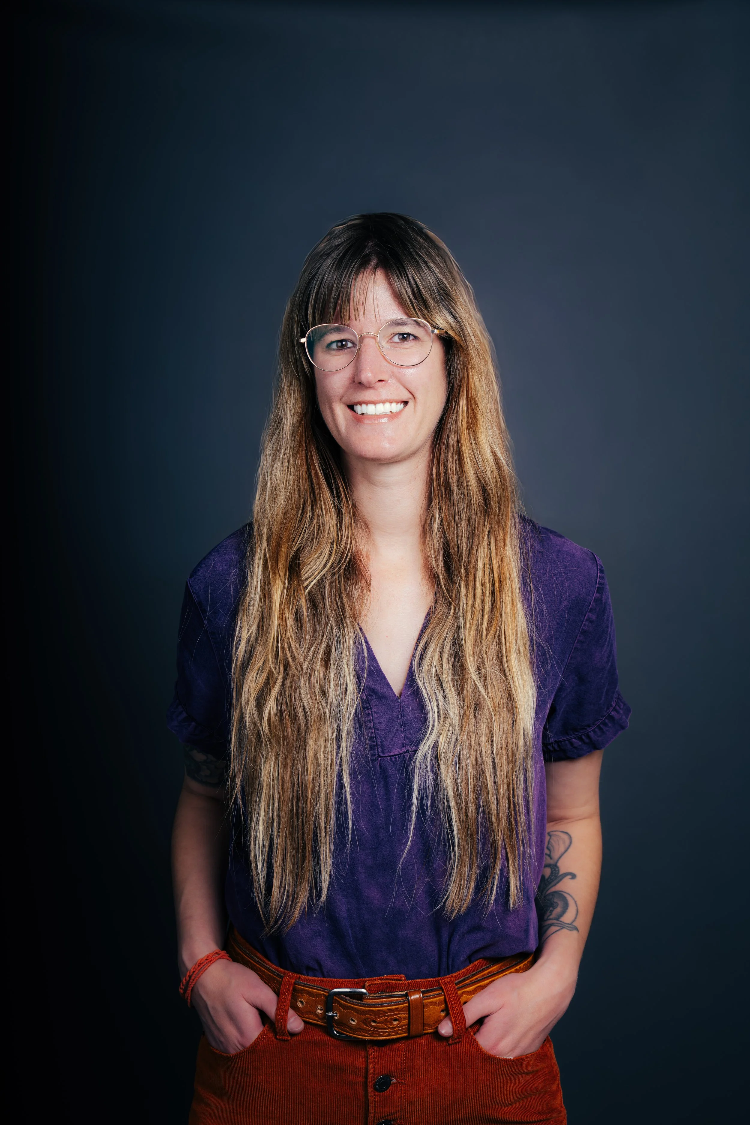A young woman with long wavy hair wearing glasses, a purple shirt, and orange pants, standing against a dark background.
