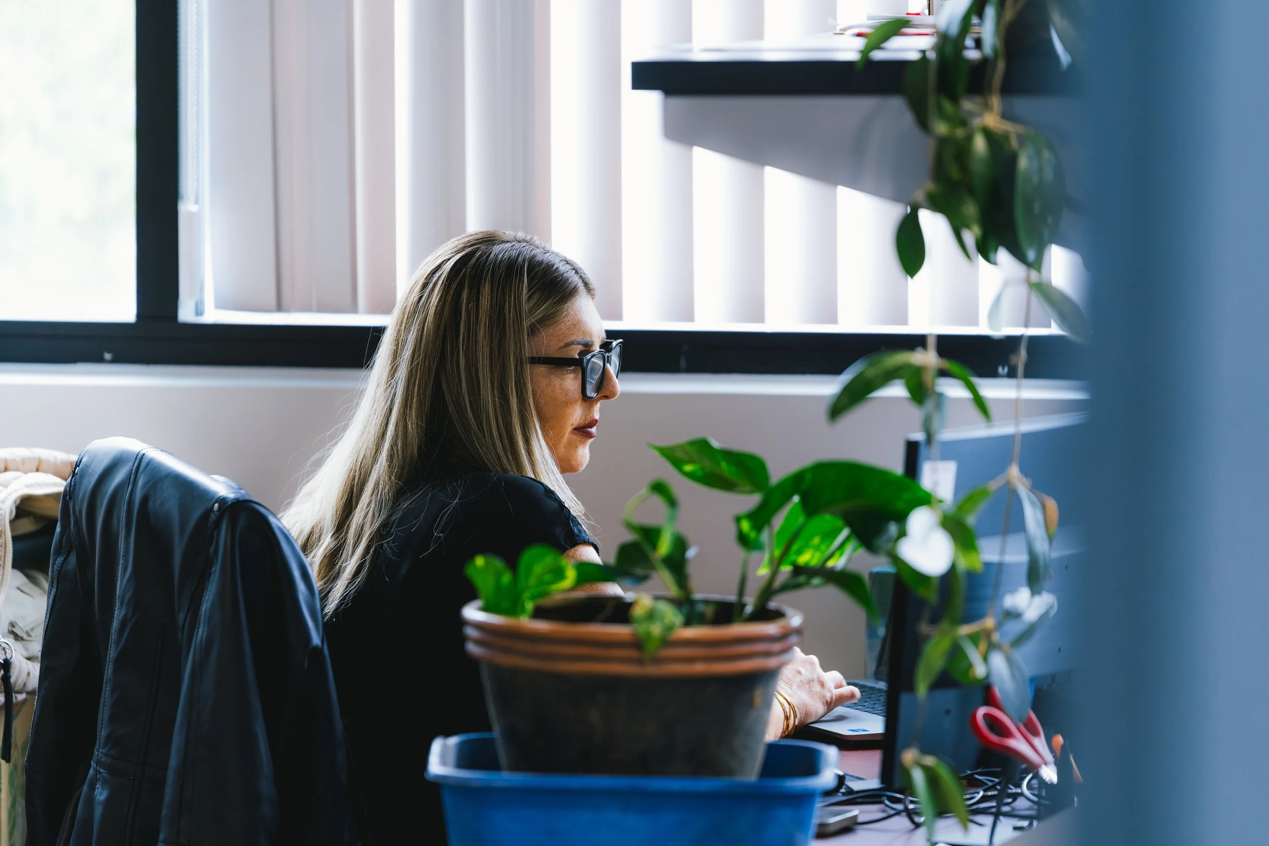 A woman with long blonde hair, glasses, and a dark top sitting at a desk with plants in the foreground, looking at her computer screen, in front of a window with vertical blinds.