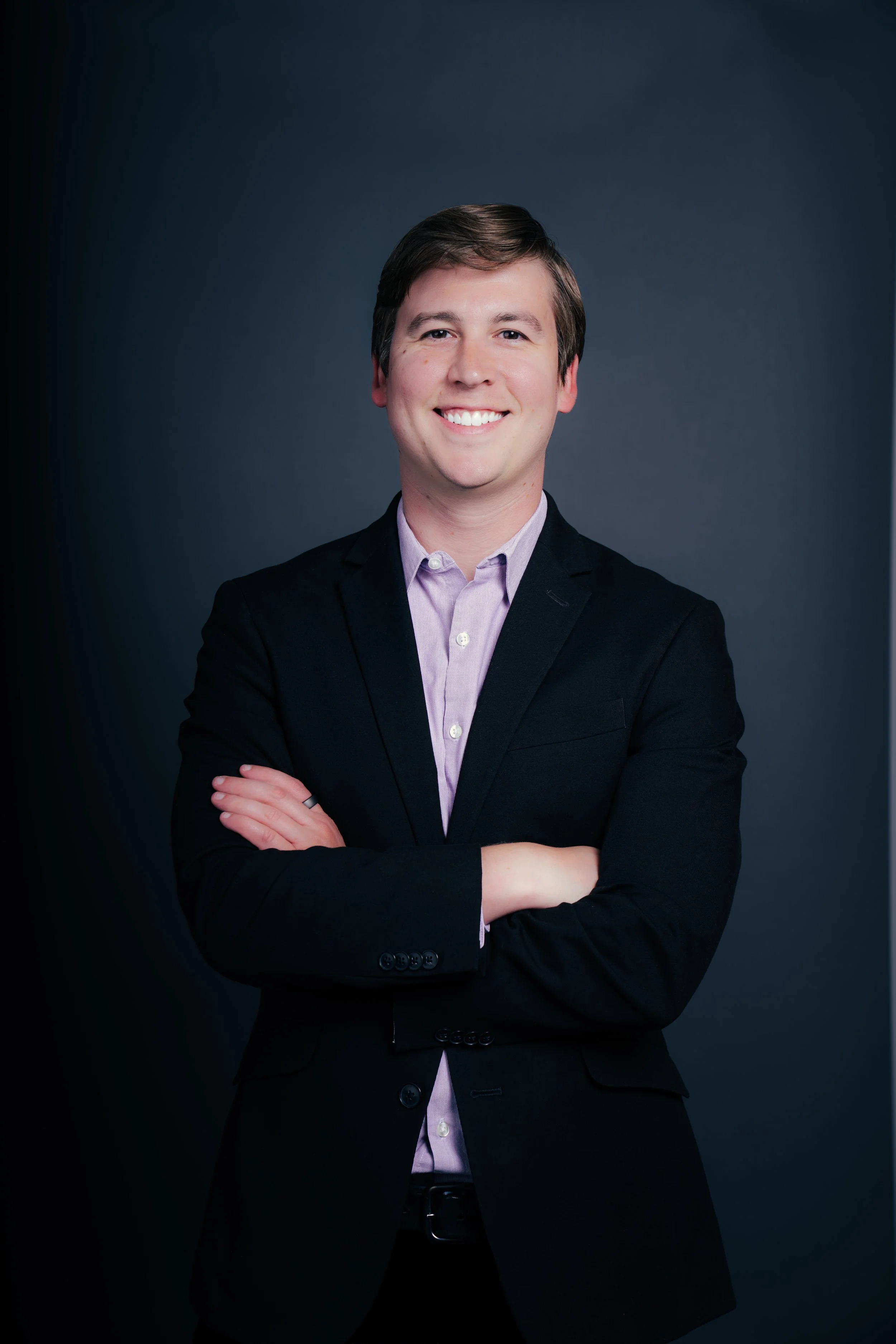 A young man with dark hair smiling, wearing a dark blazer and a light purple shirt, with arms crossed, standing against a dark background.