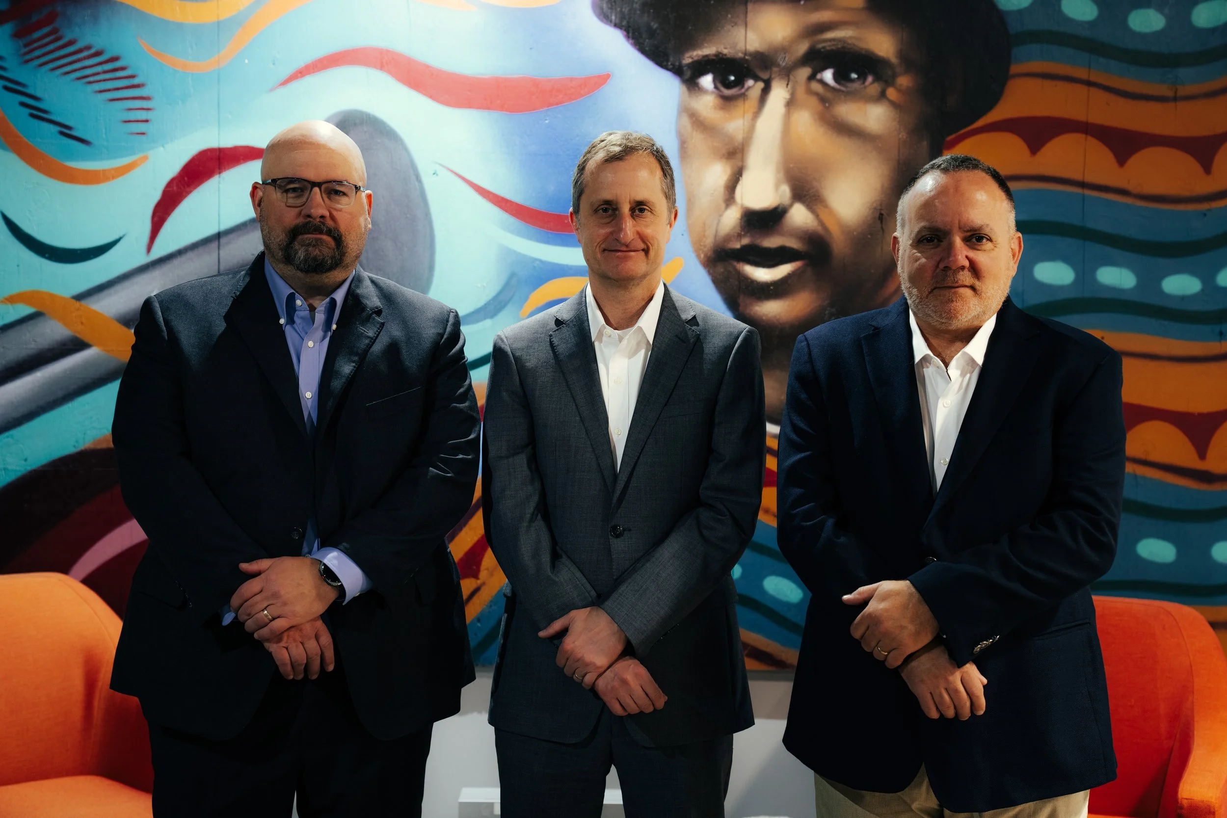 Three men in business attire standing in front of a colorful mural featuring a woman's face and swirling patterns. Two orange chairs are visible in the foreground.